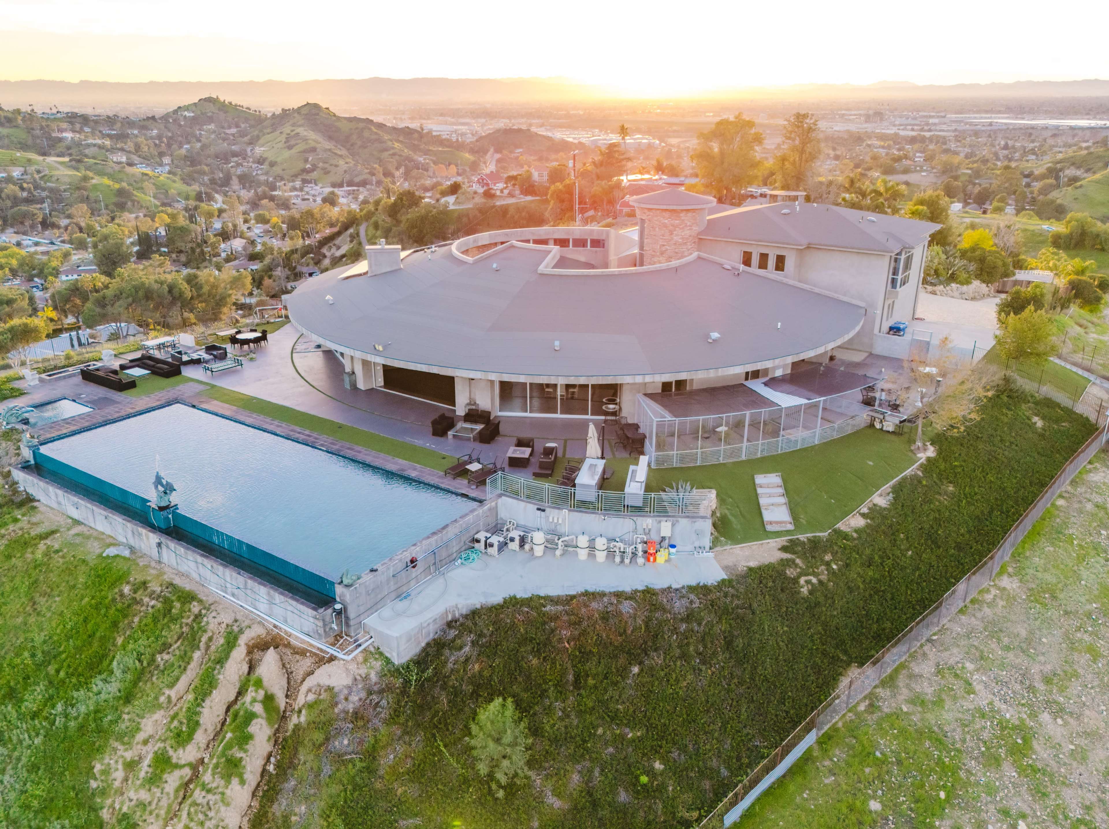The image shows a modern circular house with a large swimming pool and landscaped outdoor area, situated on a hillside during sunset.