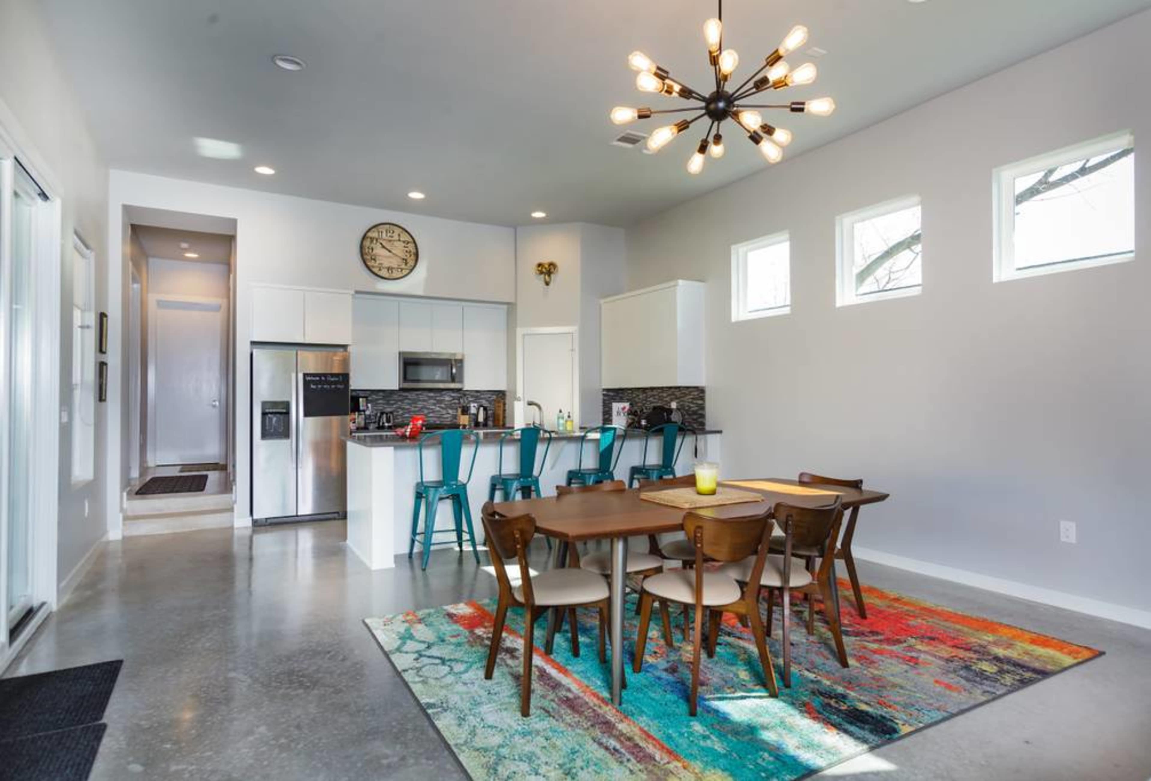 A modern kitchen and dining area with a large table, colorful rug, and contemporary lighting fixtures.
