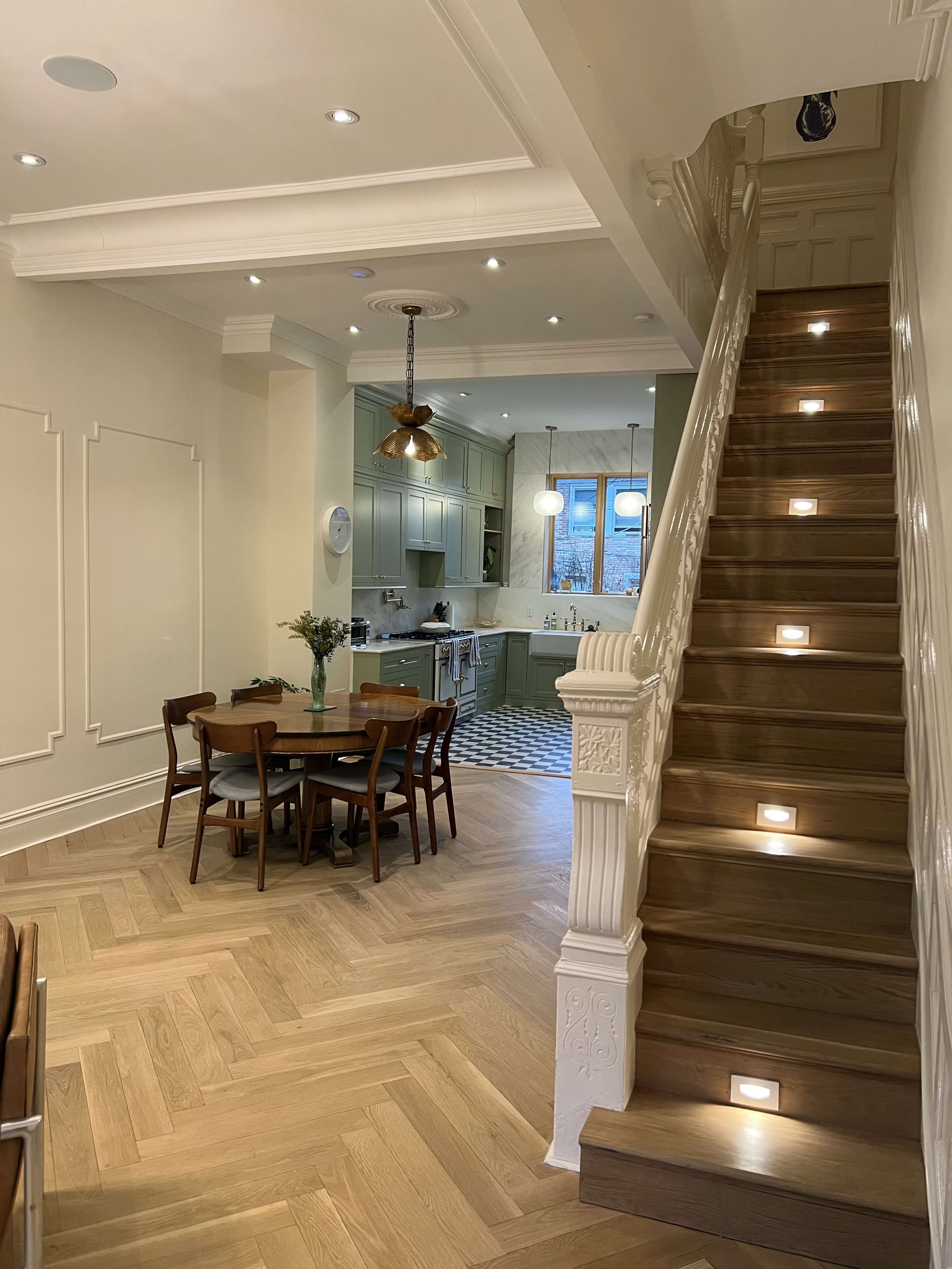A well-lit interior space featuring a staircase with recessed lighting, leading to a kitchen and dining area furnished with a round table and wooden chairs.