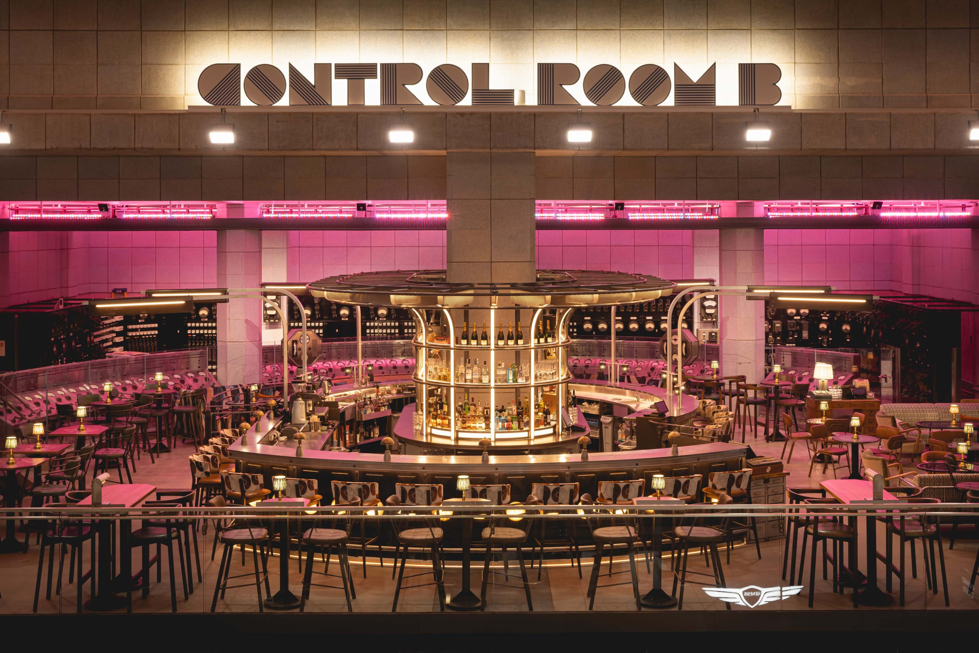 A bar area named "CONTROL ROOM B," featuring a circular bar surrounded by high stools, with pink lighting illuminating the space.