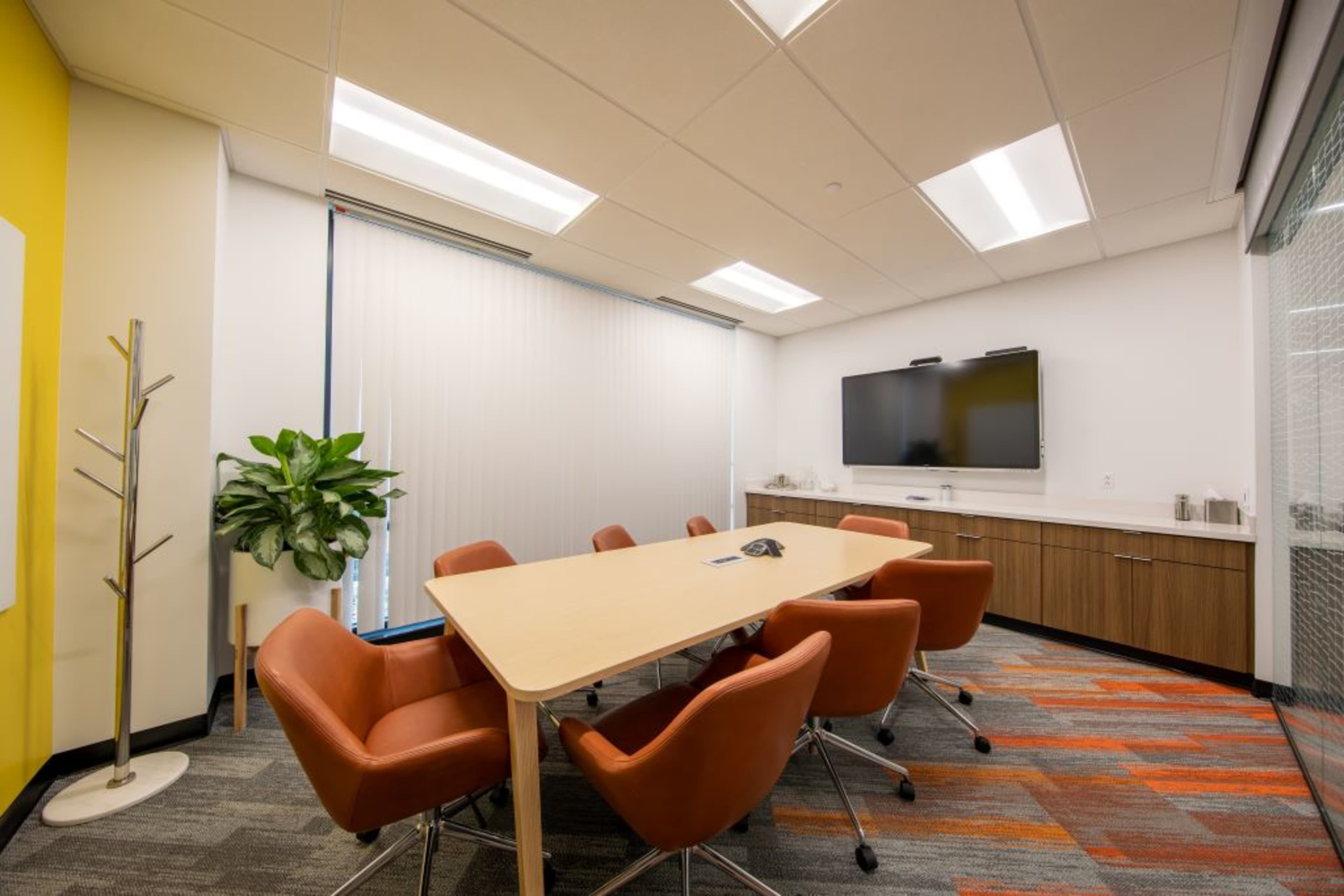 A conference room features a large table surrounded by orange chairs, a television mounted on the wall, and a potted plant in the corner.