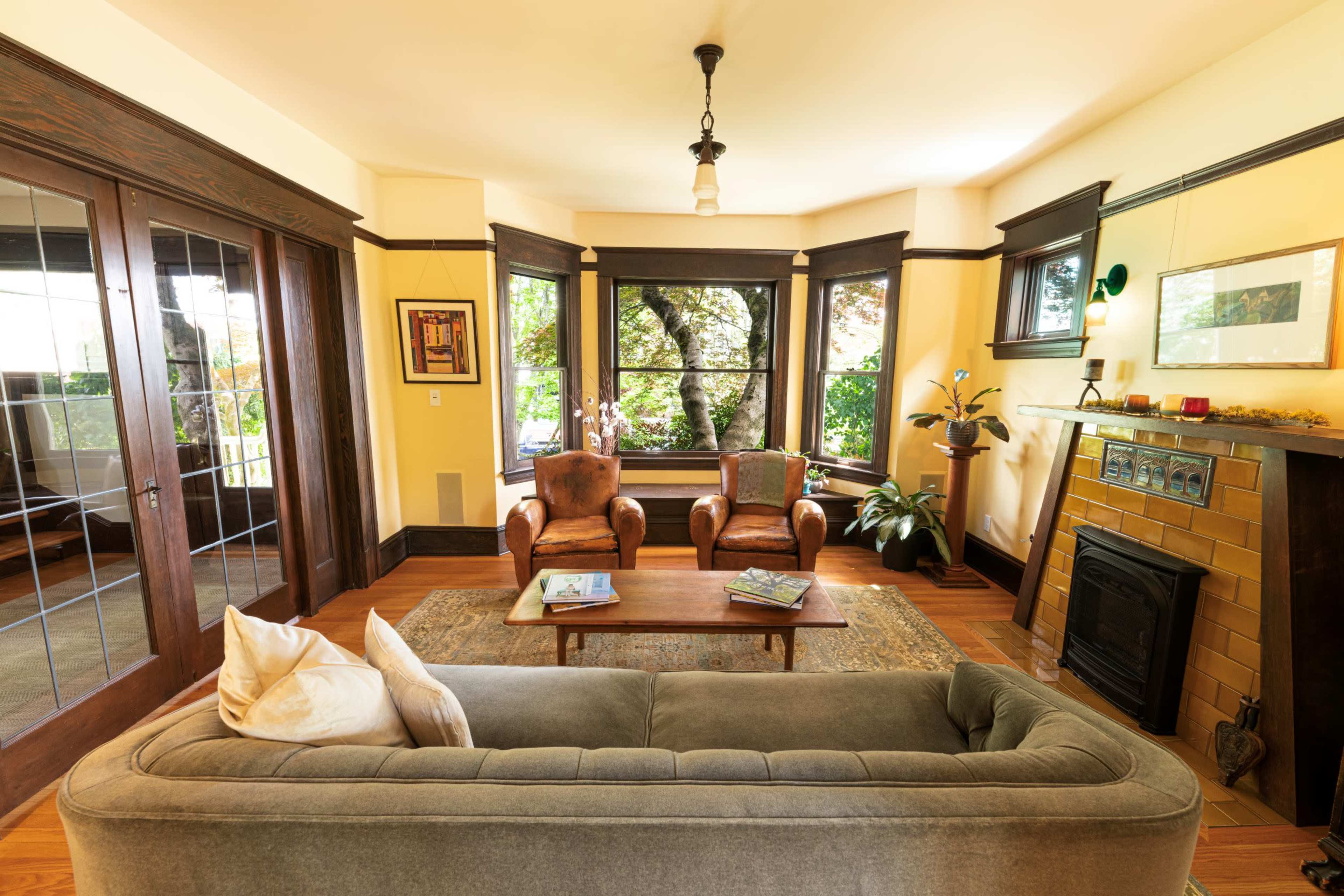 A cozy living room with two armchairs, a coffee table, and a fireplace, surrounded by large windows that offer a view of outdoor greenery.
