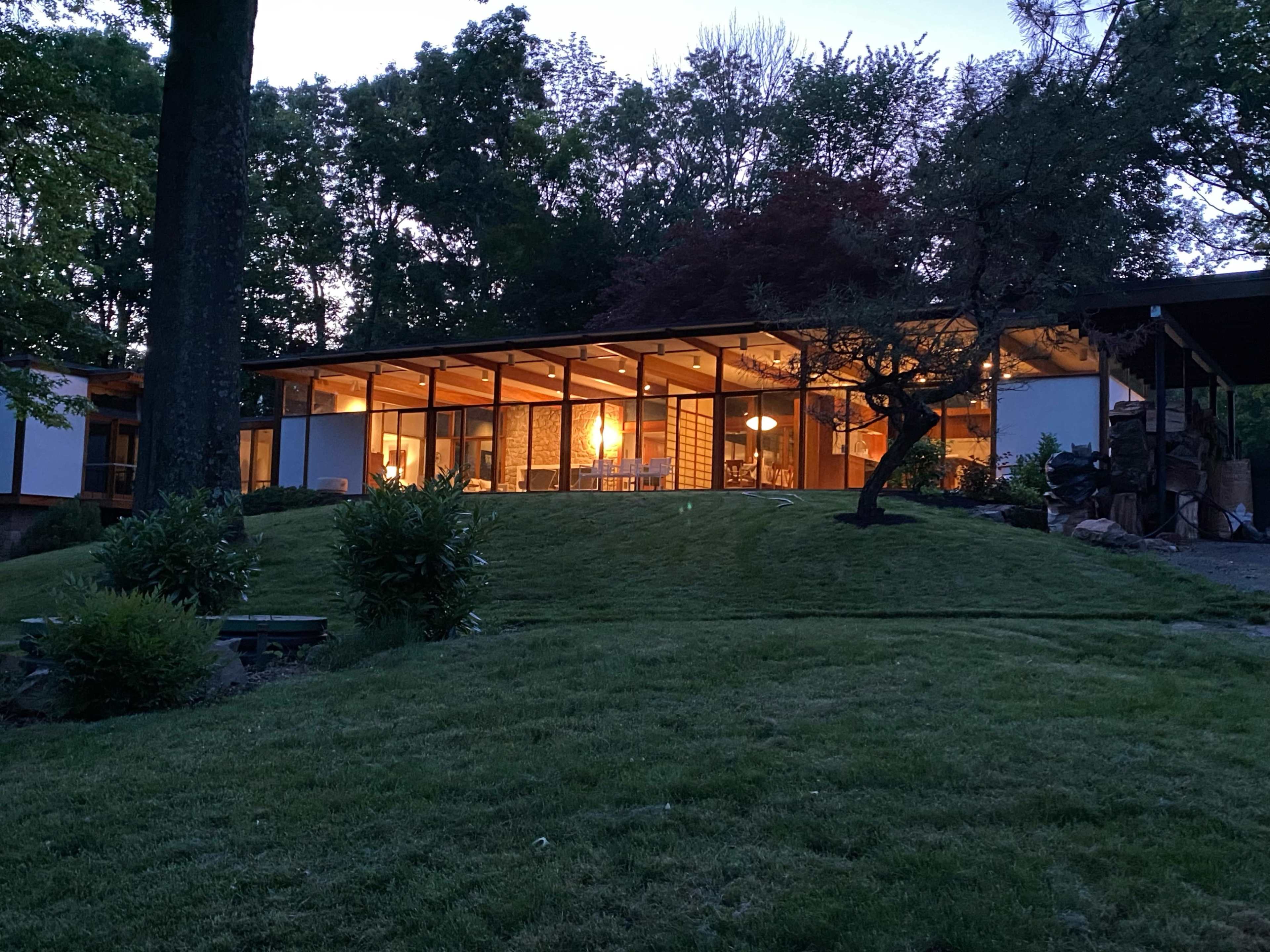 A modern house with large windows and a wooden structure is set on a landscaped hill surrounded by trees at dusk.