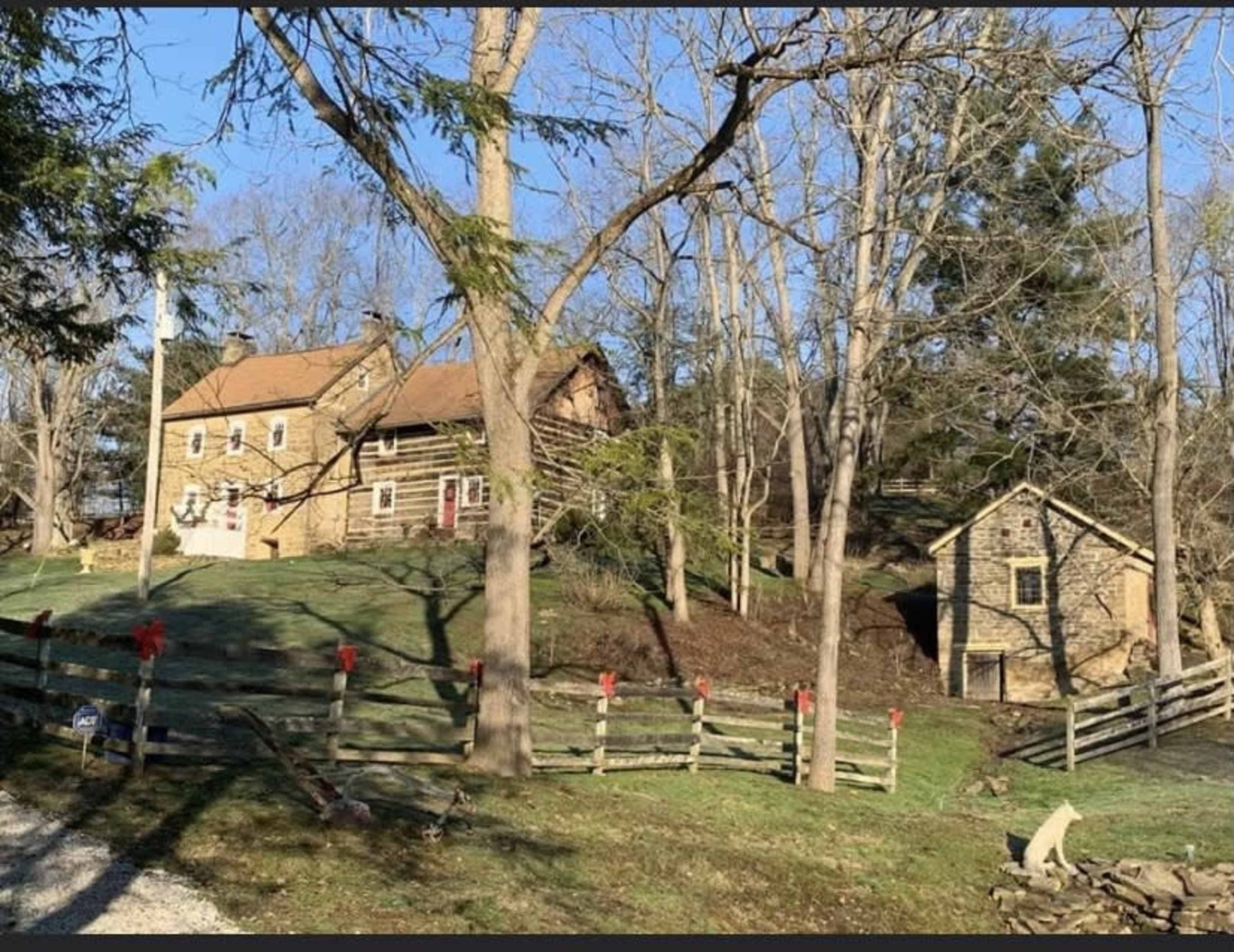 A brick farmhouse surrounded by trees and a wooden fence, with a smaller stone building nearby and holiday decorations on the fence.