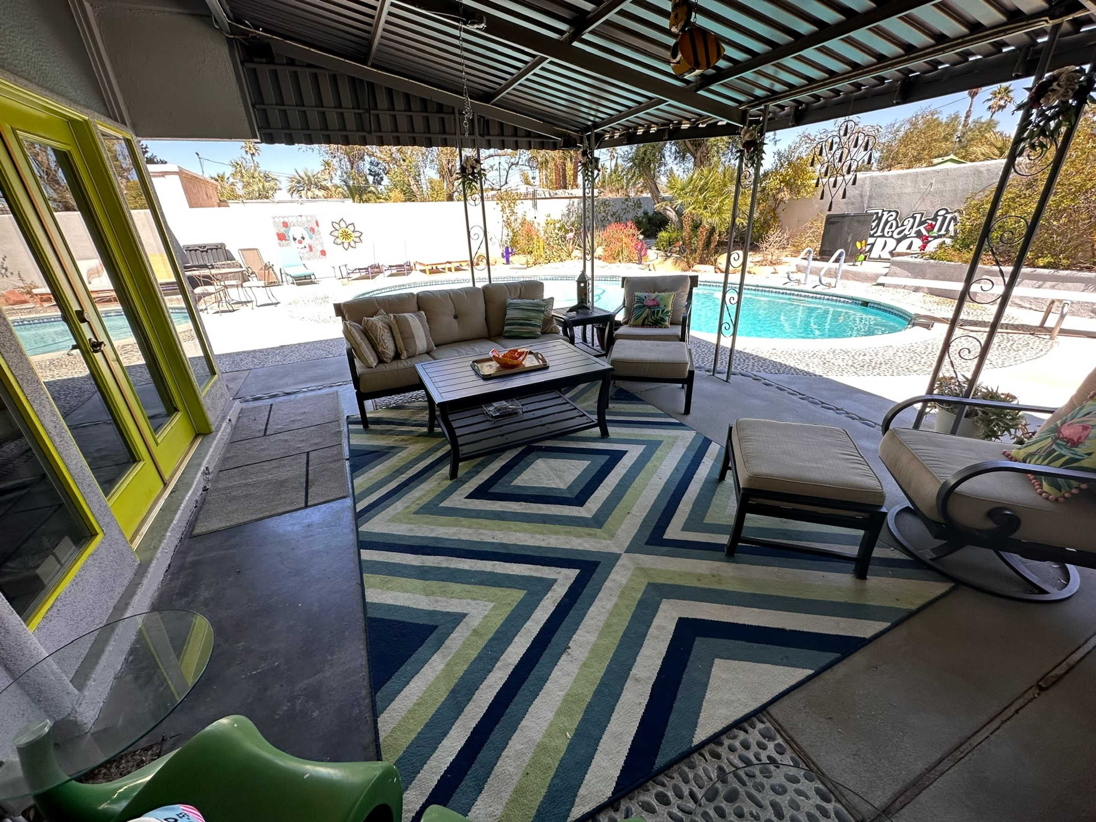 A covered patio area with seating, a patterned rug, and a view of a swimming pool surrounded by landscaping.
