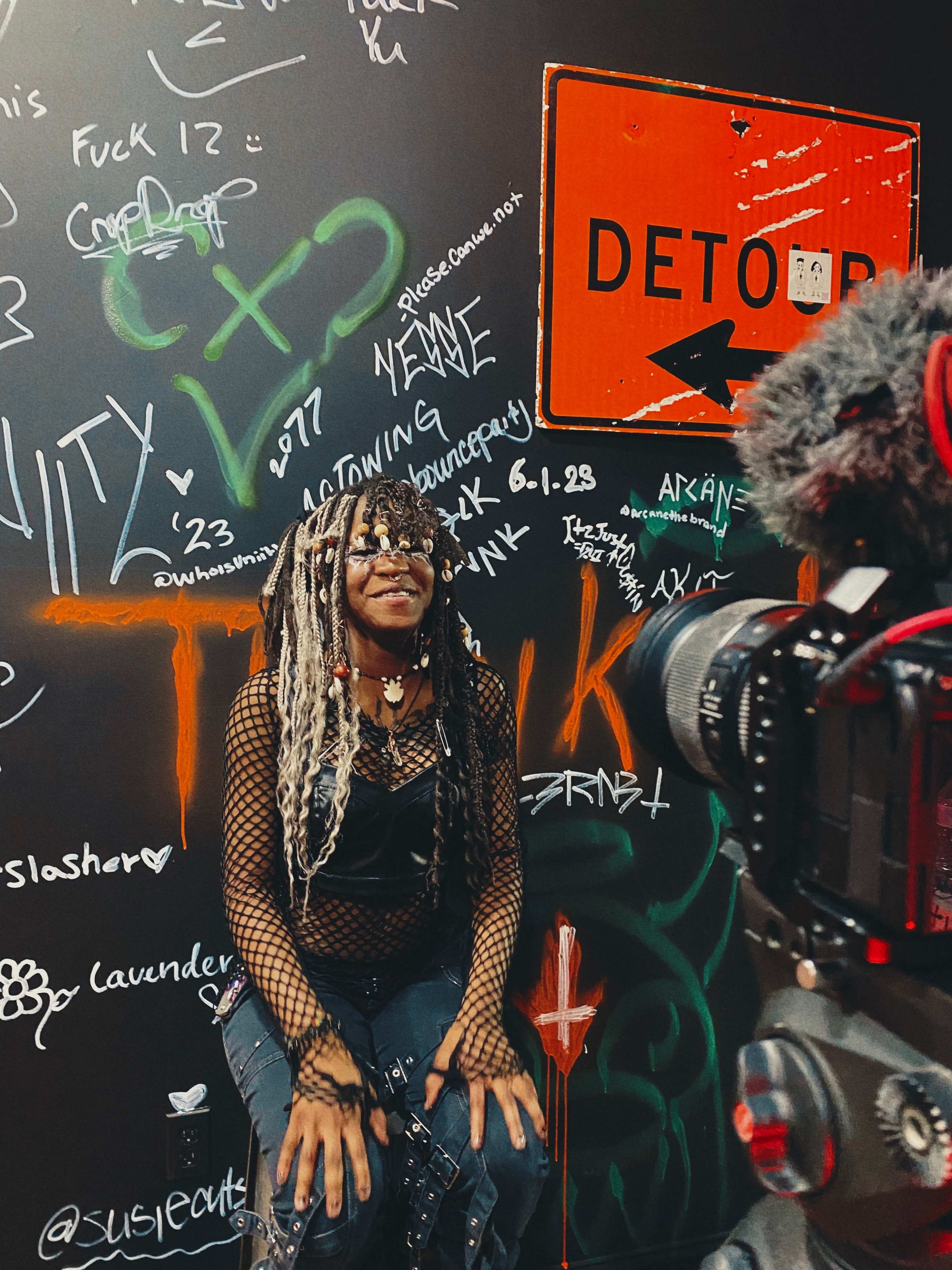 A person with textured hair and a distinctive outfit sits in front of a graffiti-covered wall while being filmed by a camera.