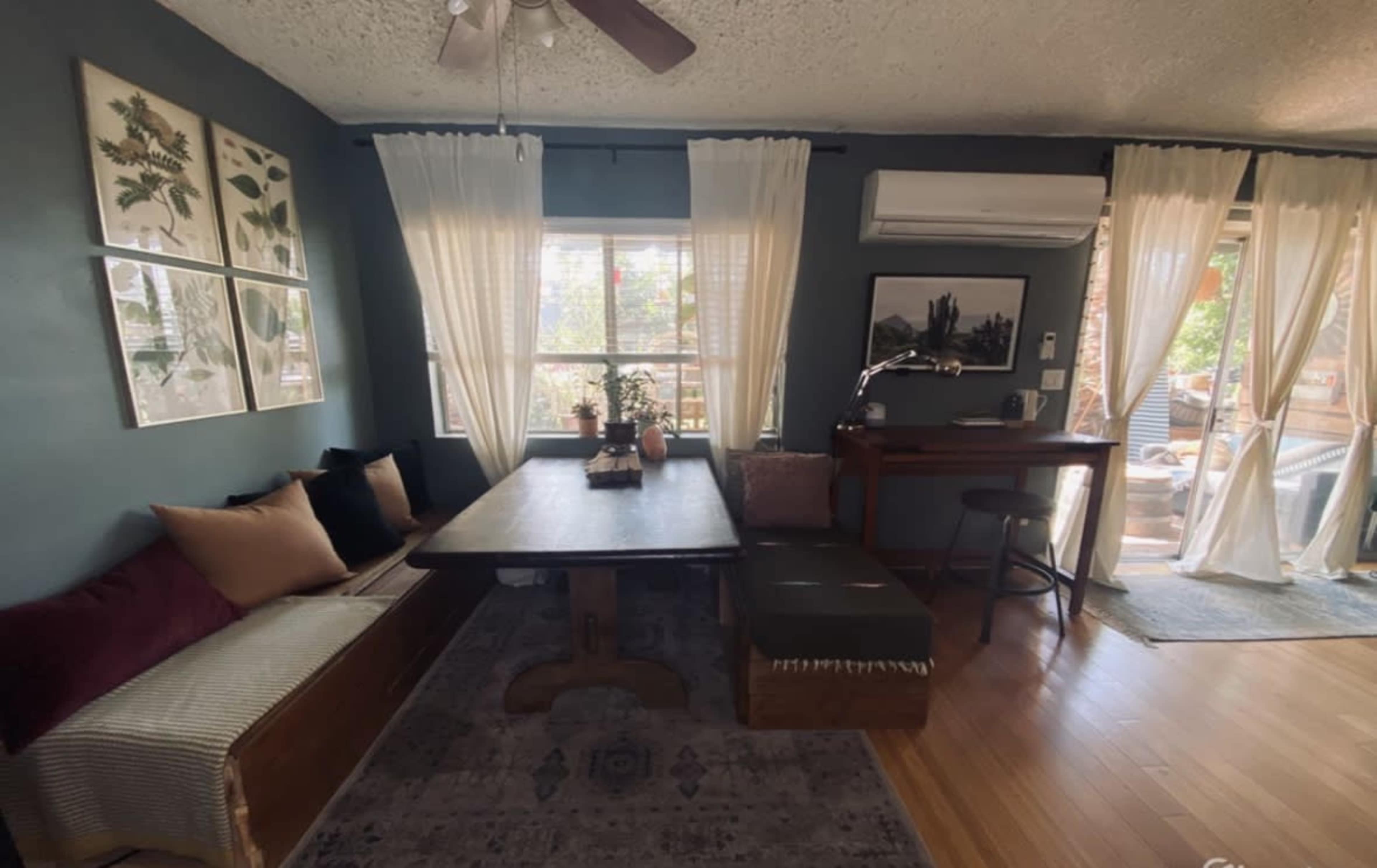 A cozy dining area with a wooden table, a window with sheer curtains, and a small bar-style desk along the wall.