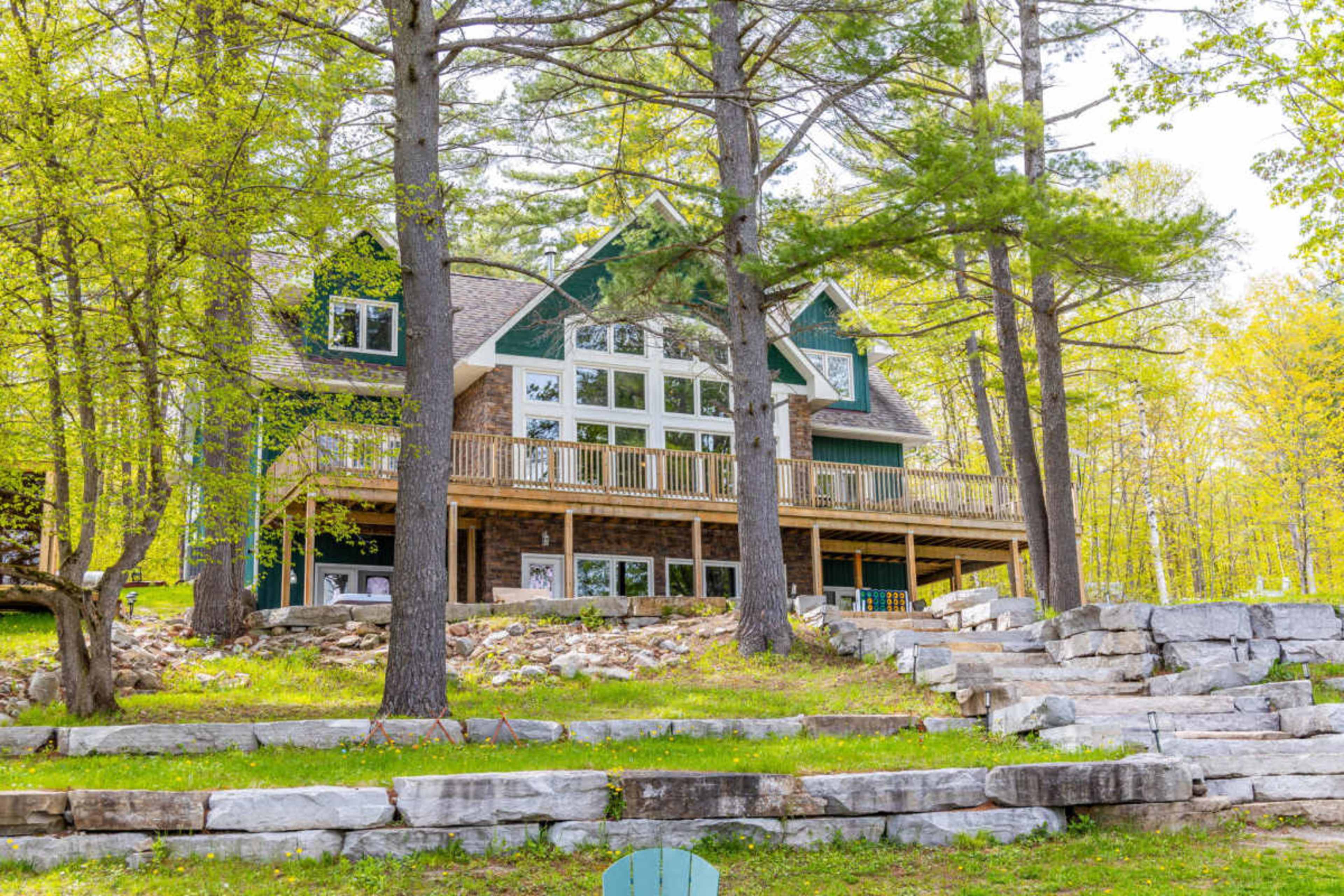 A large, multi-story house with a stone facade and expansive deck is nestled among tall trees and landscaped stone steps.