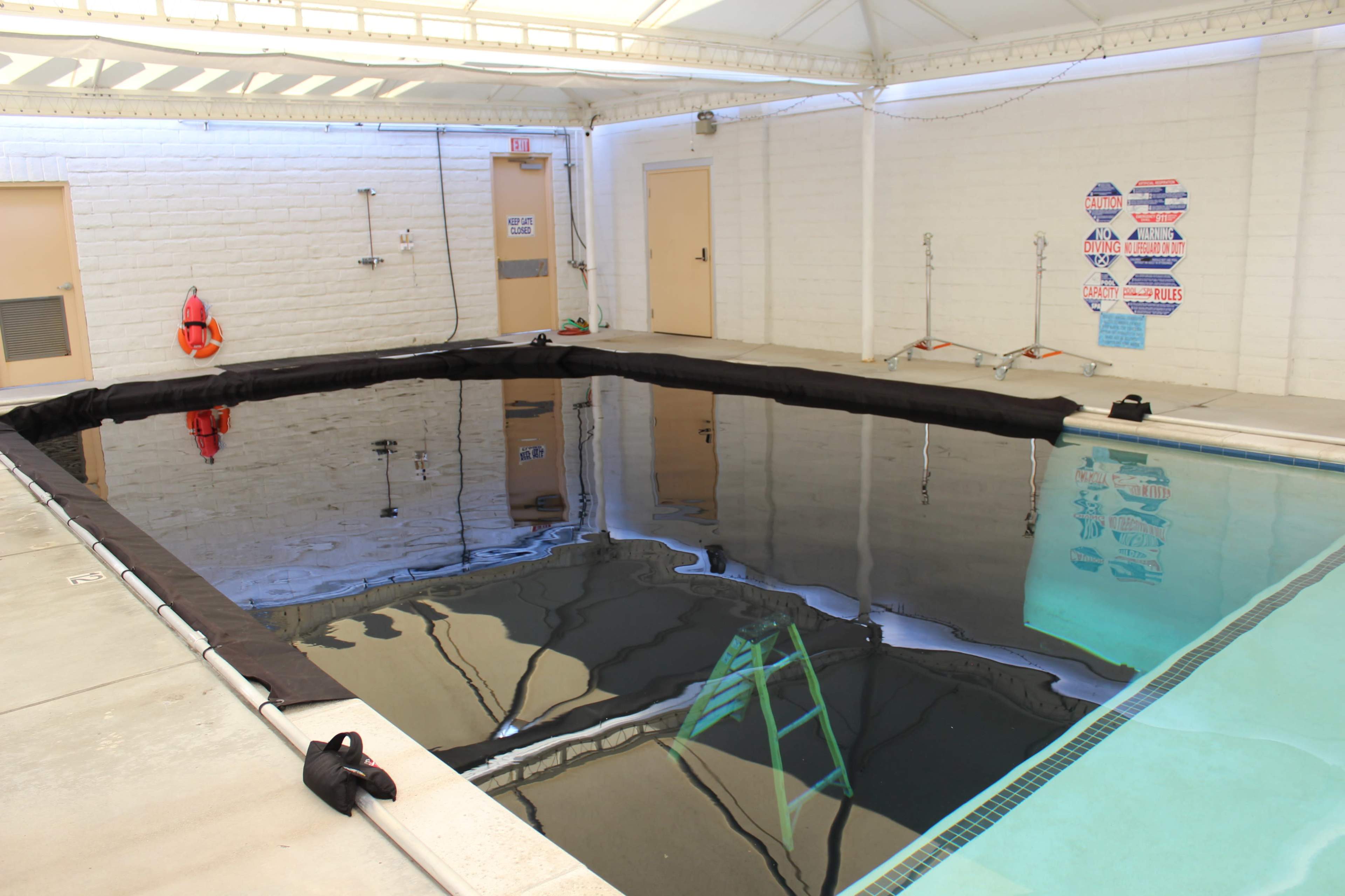 An indoor swimming pool is partially covered with a black tarp, with a ladder leaning against the edge.
