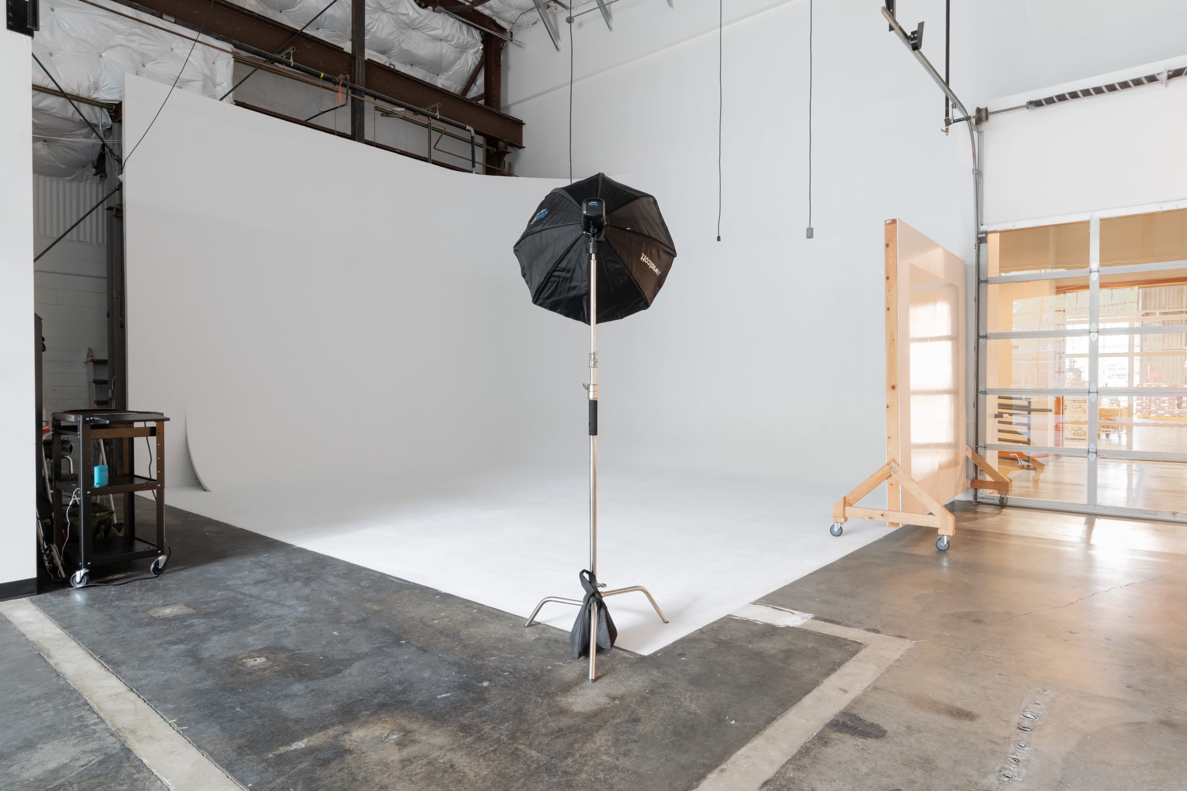 The image shows a photography studio featuring a white backdrop, a black umbrella light stand, and a large, empty space with concrete flooring.
