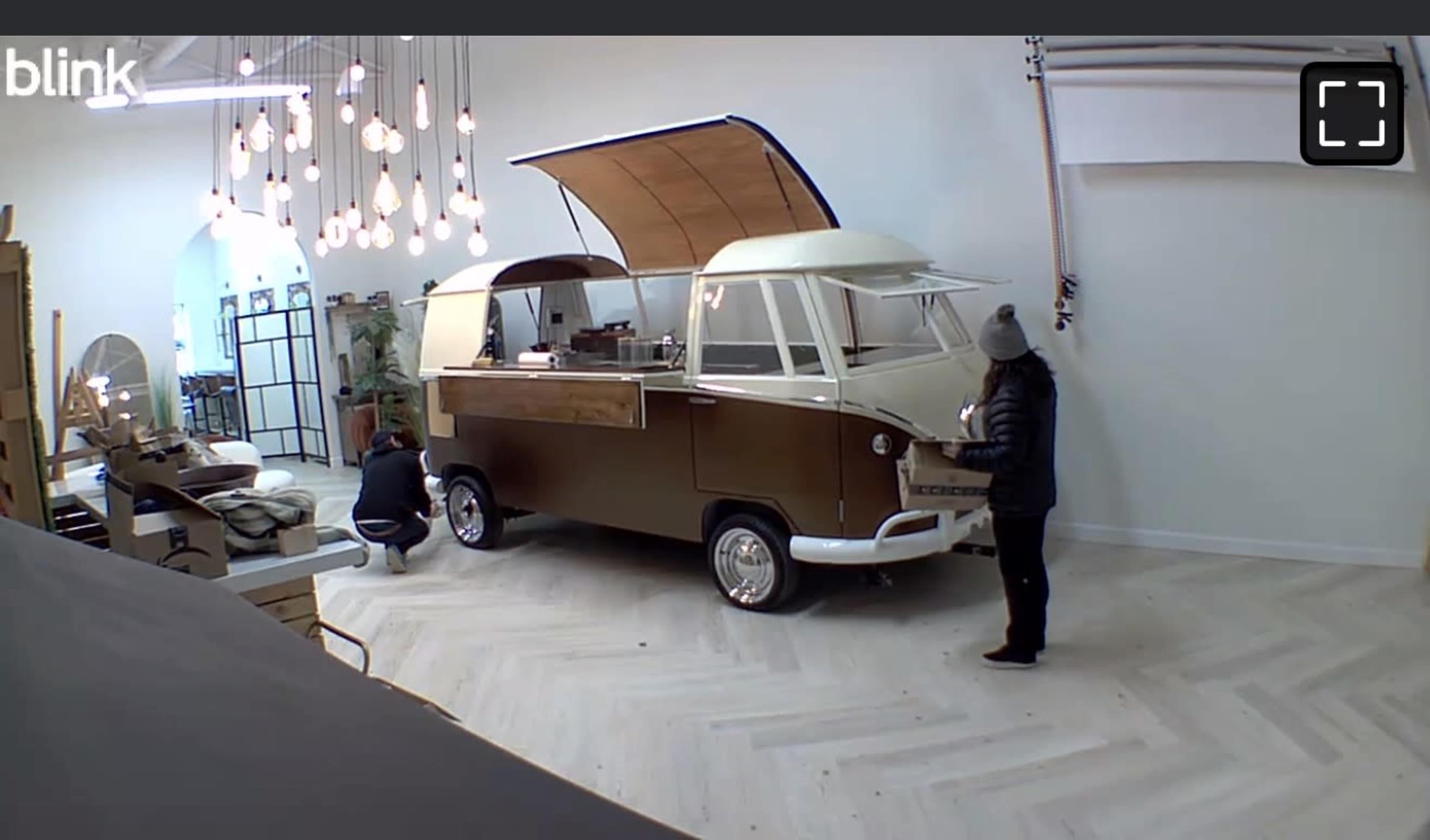 Two people work on a vintage-style food truck inside a well-lit, modern interior space with wooden flooring and hanging light fixtures.