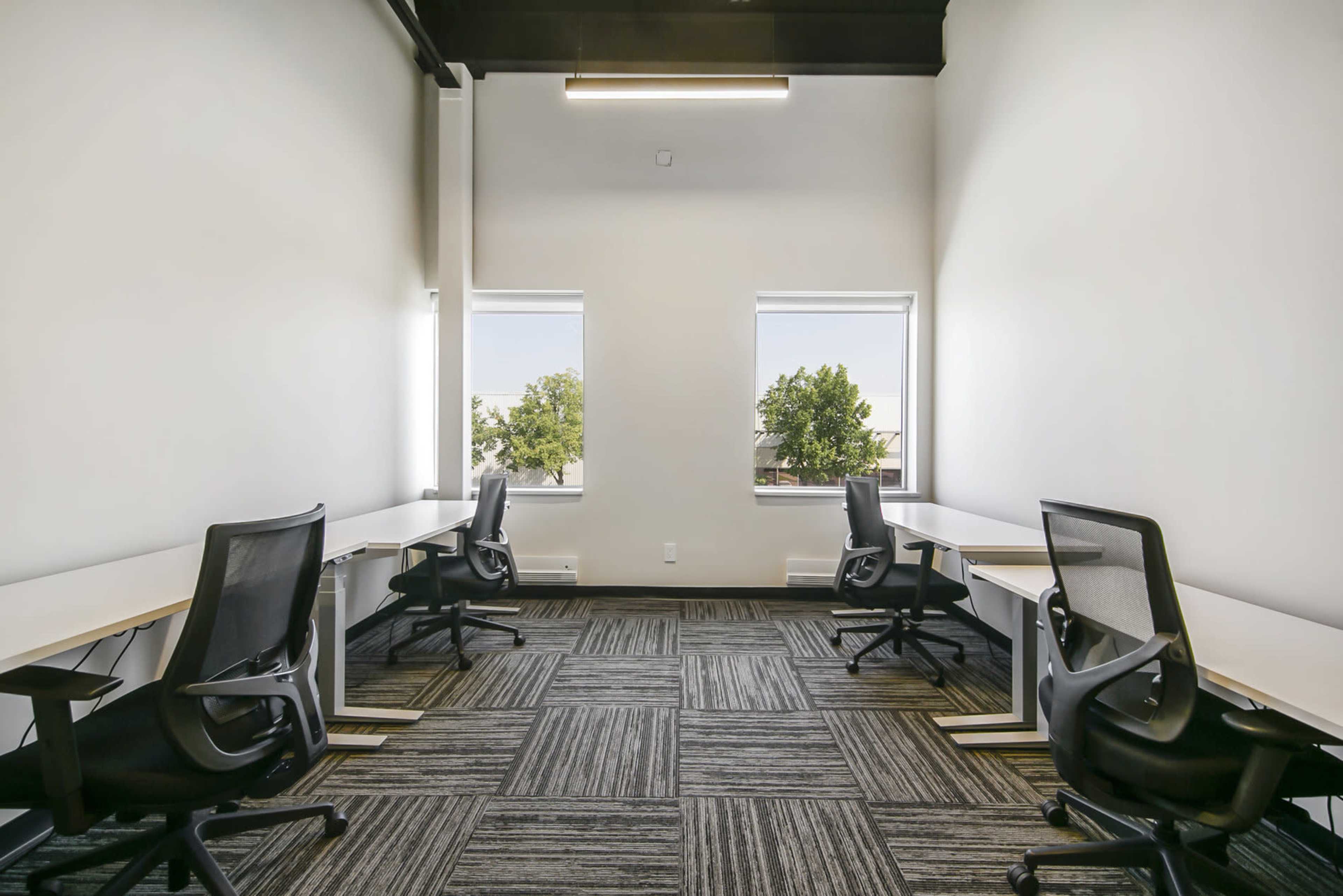The image shows a small office space with four black ergonomic chairs and white desks arranged along two walls, with windows letting in natural light.