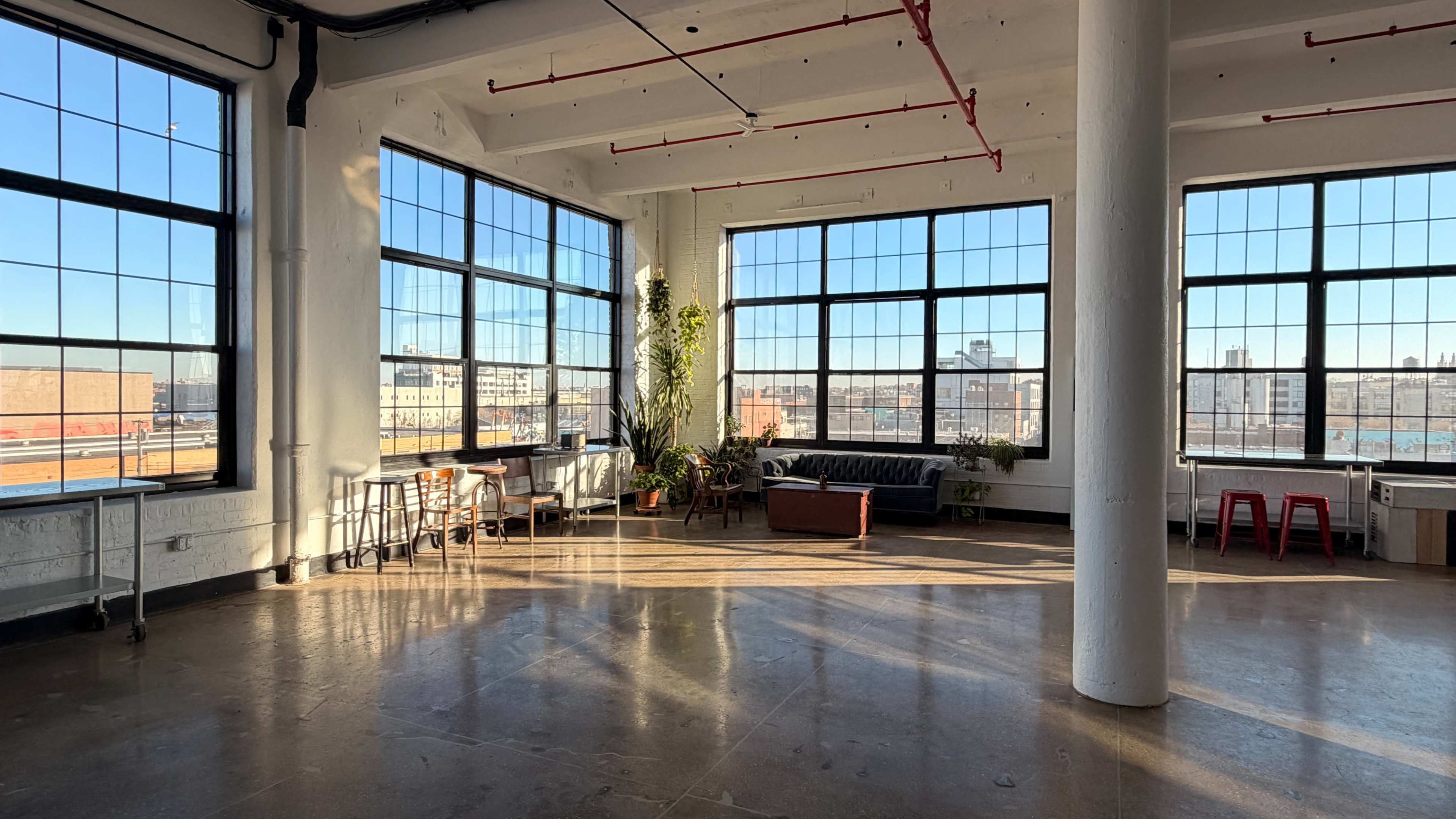 The image shows a spacious, well-lit room with large windows, modern furniture, and plants, overlooking an urban landscape.