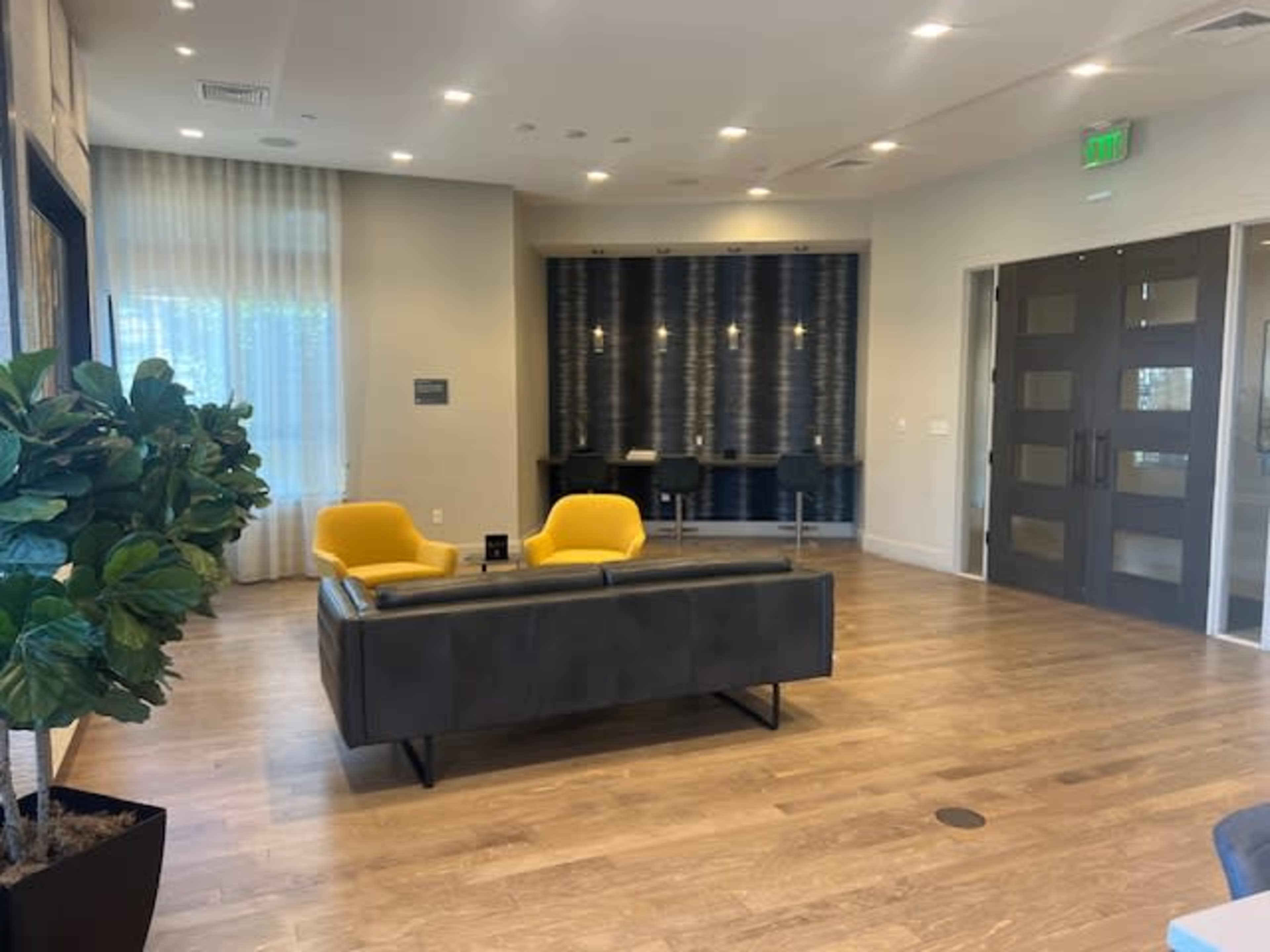 A spacious lobby area featuring a gray sofa and two yellow chairs, with a plant in the corner and a reception desk in the background.