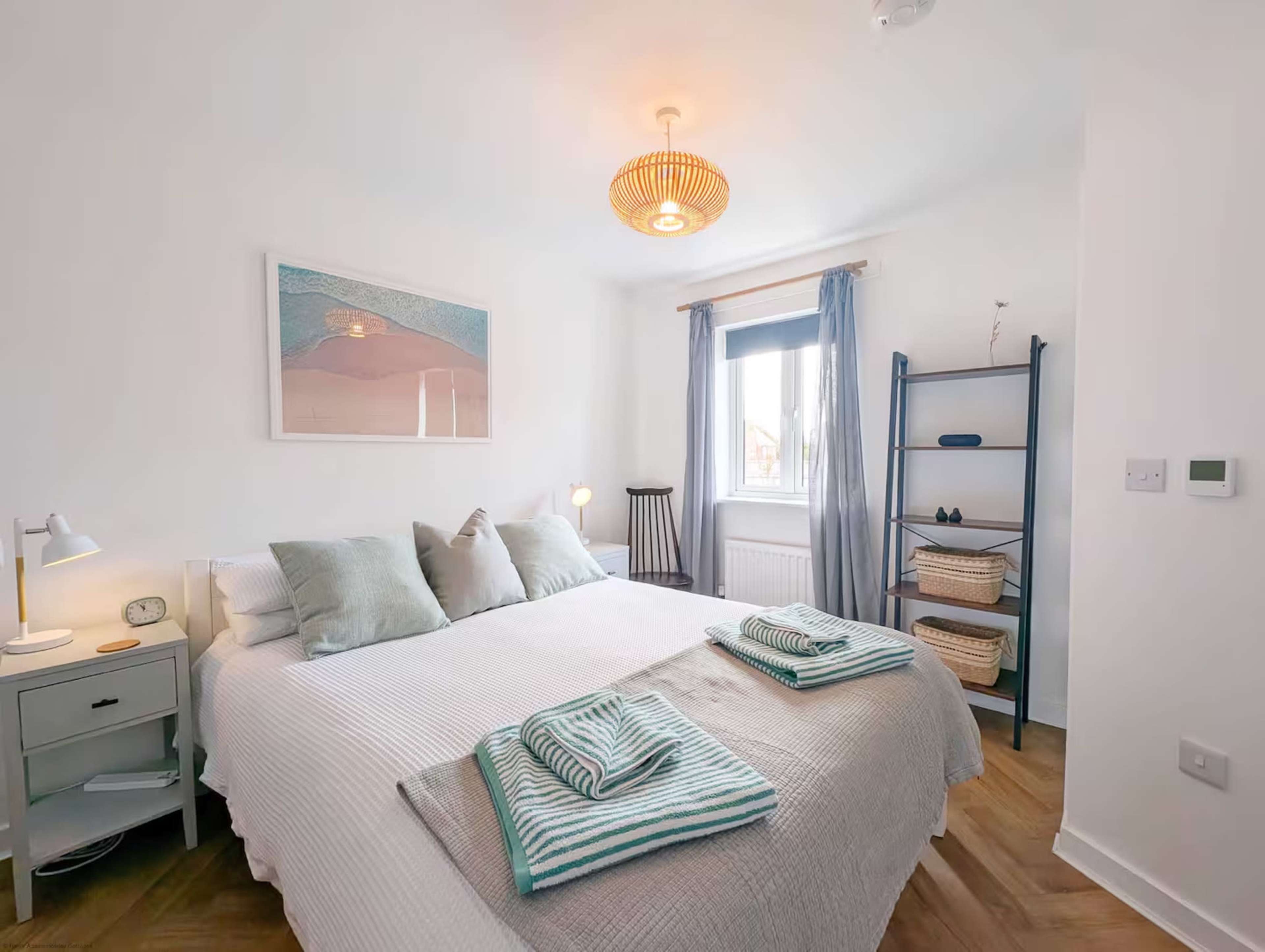 The image shows a neatly arranged bedroom featuring a large bed with light-colored bedding, two bedside tables, and a decorative rug, complemented by natural light from a window.