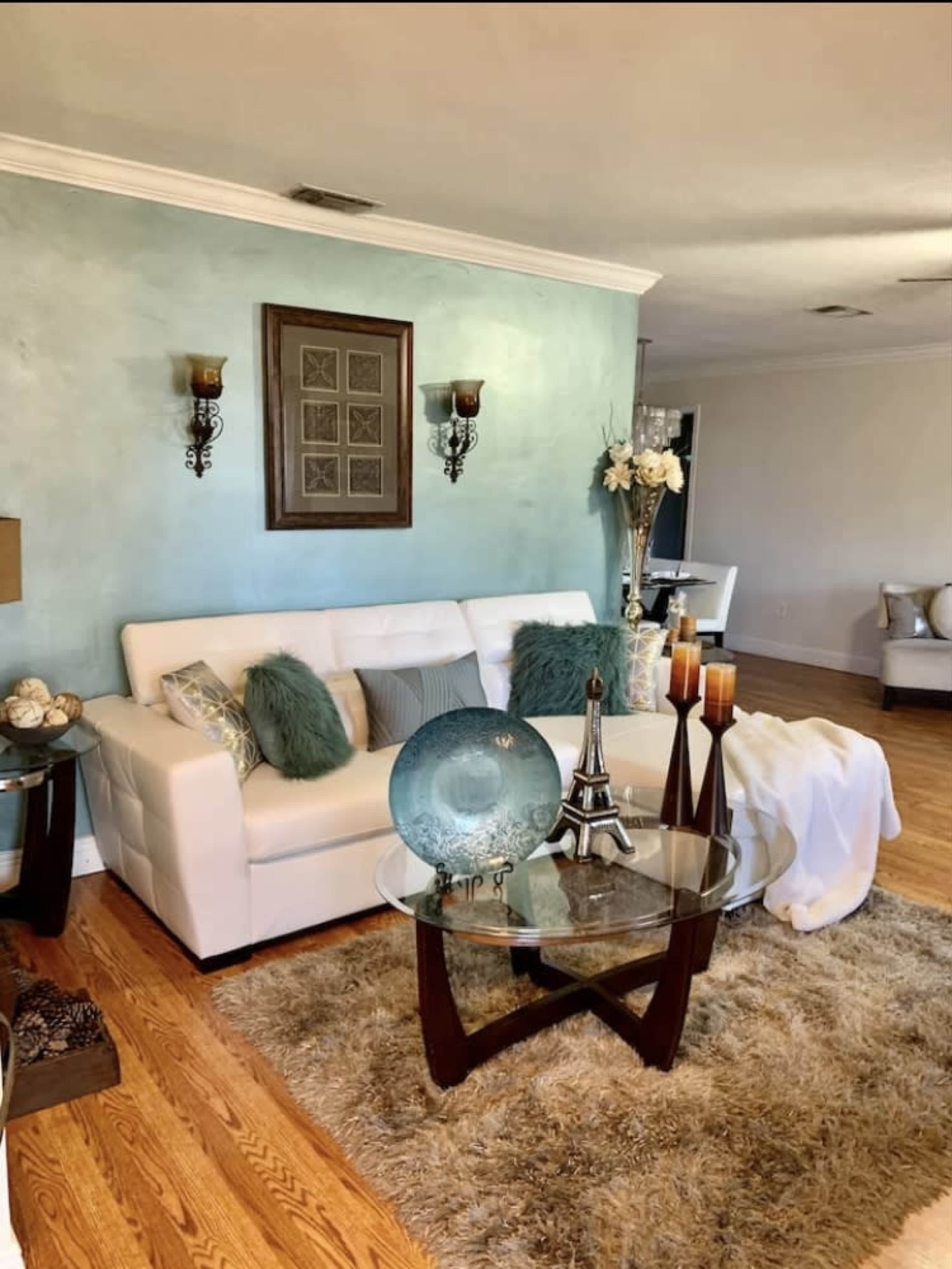 The living room features a white sofa with decorative pillows, a glass coffee table, and a textured area rug, set against a light blue wall.