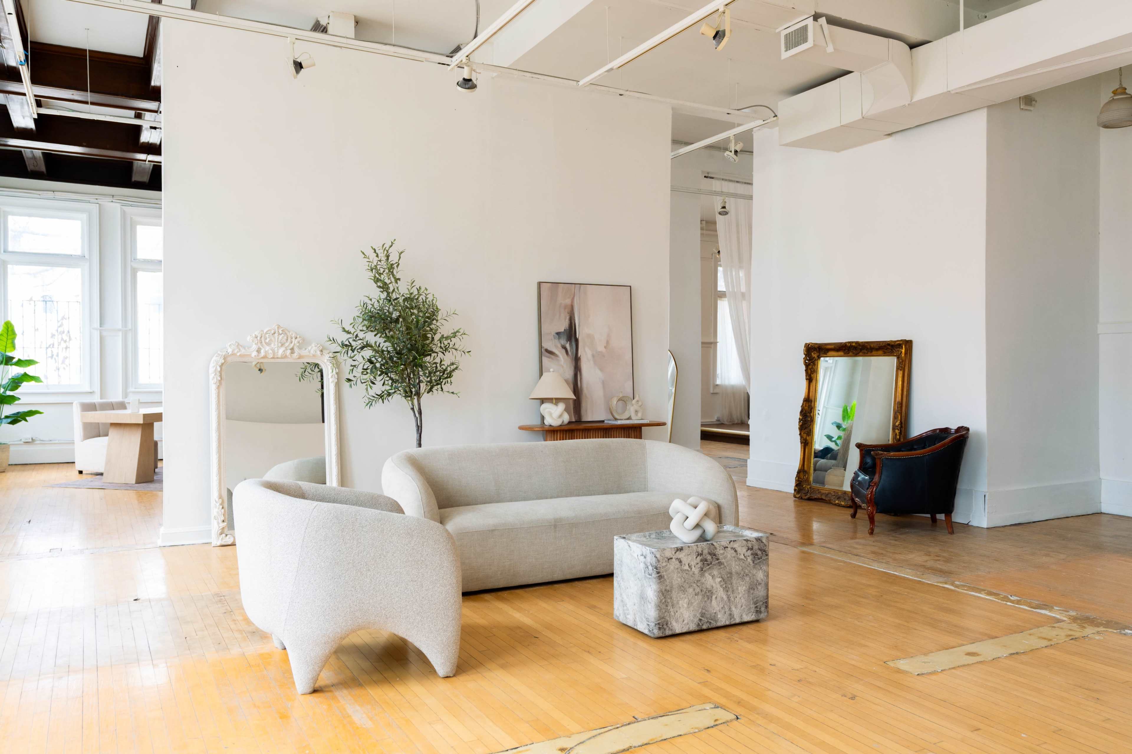 The image shows a modern, minimalist living space featuring a curved sofa, a marble coffee table, and artwork on the walls, with ample natural light flooding the room.
