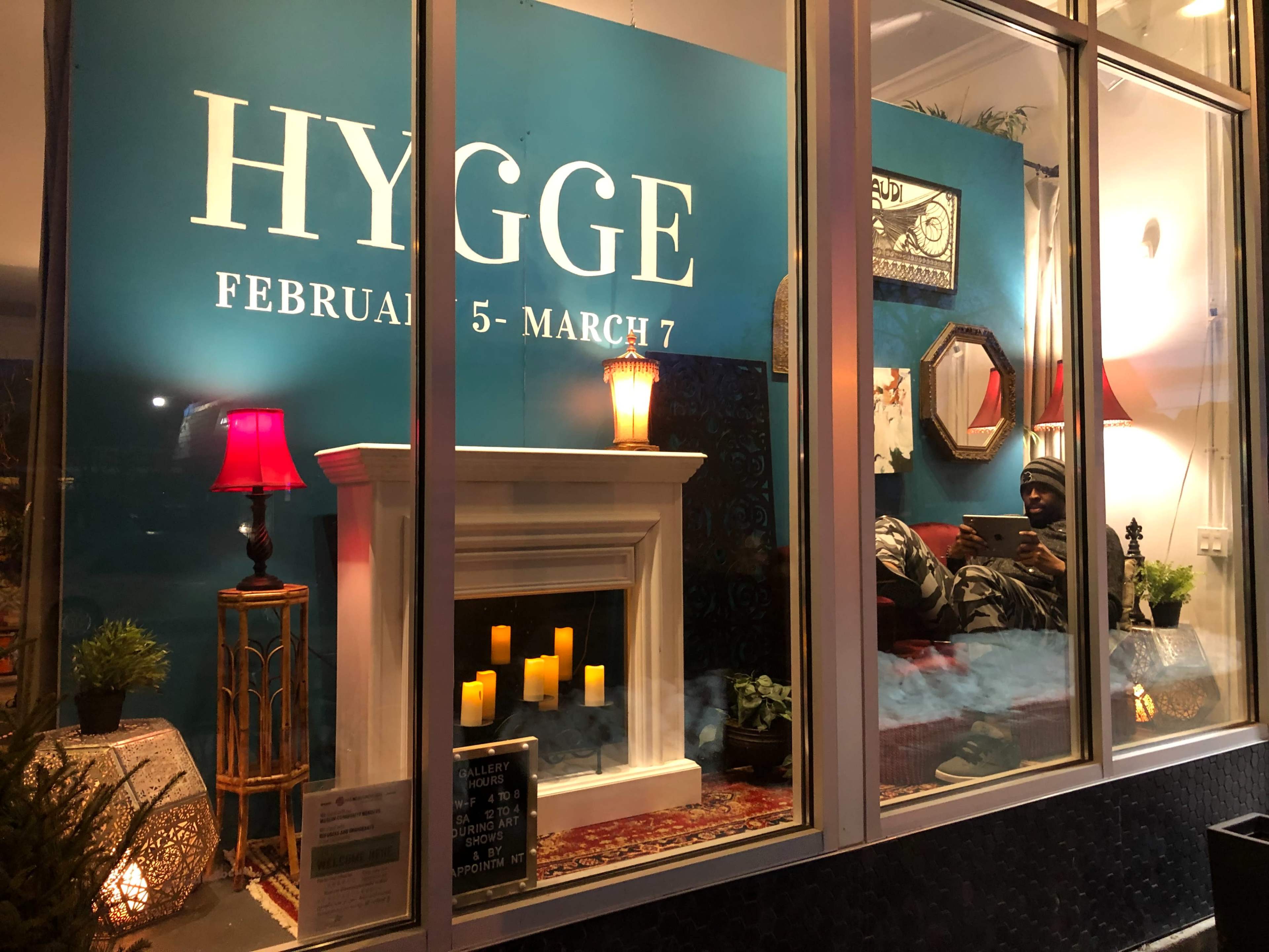 A cozy storefront display features a fireplace, candles, plants, and a person seated comfortably in a well-lit area.