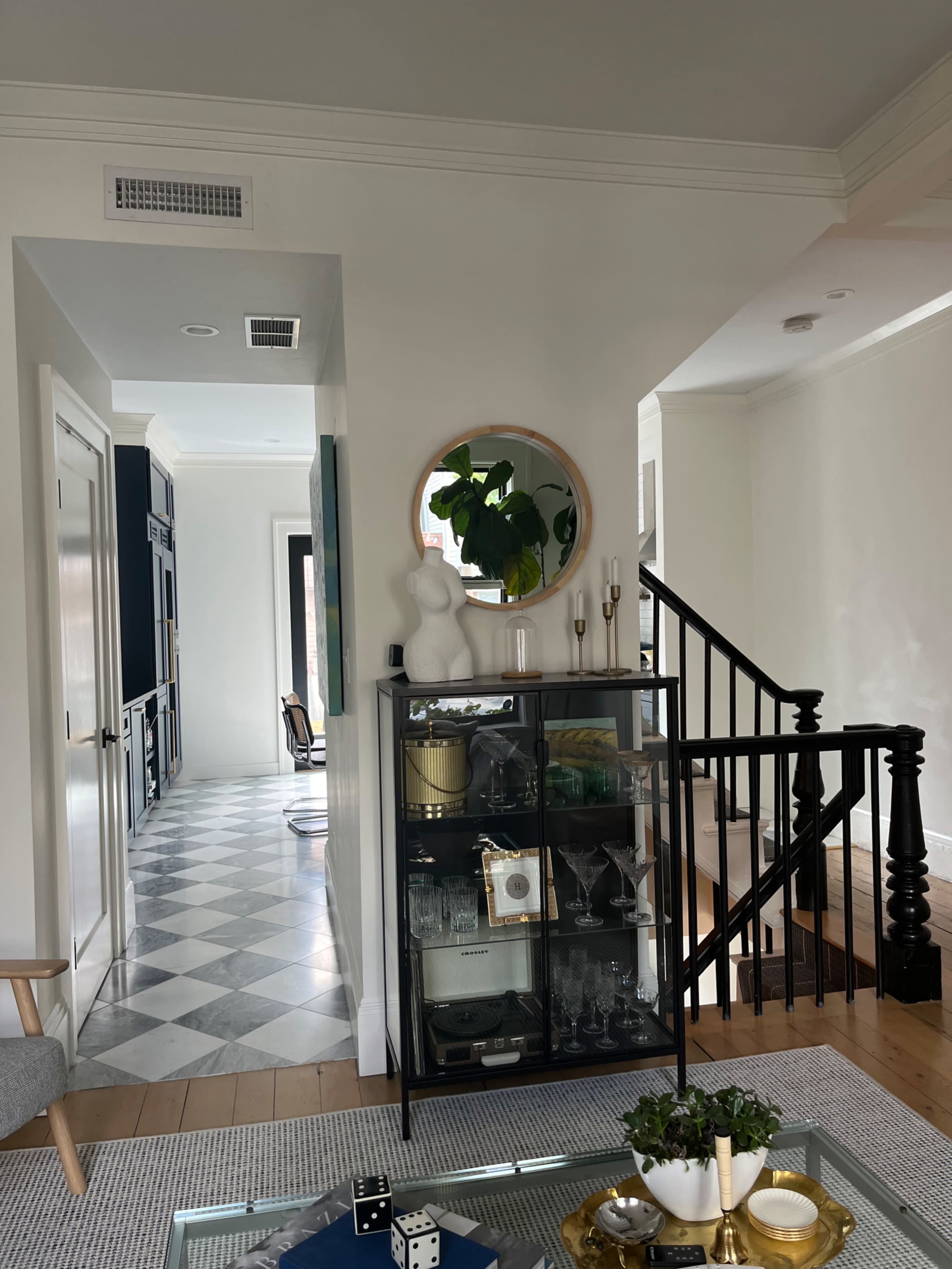 A glass-front cabinet displaying various glassware is positioned near a staircase in a well-lit hallway with patterned flooring.