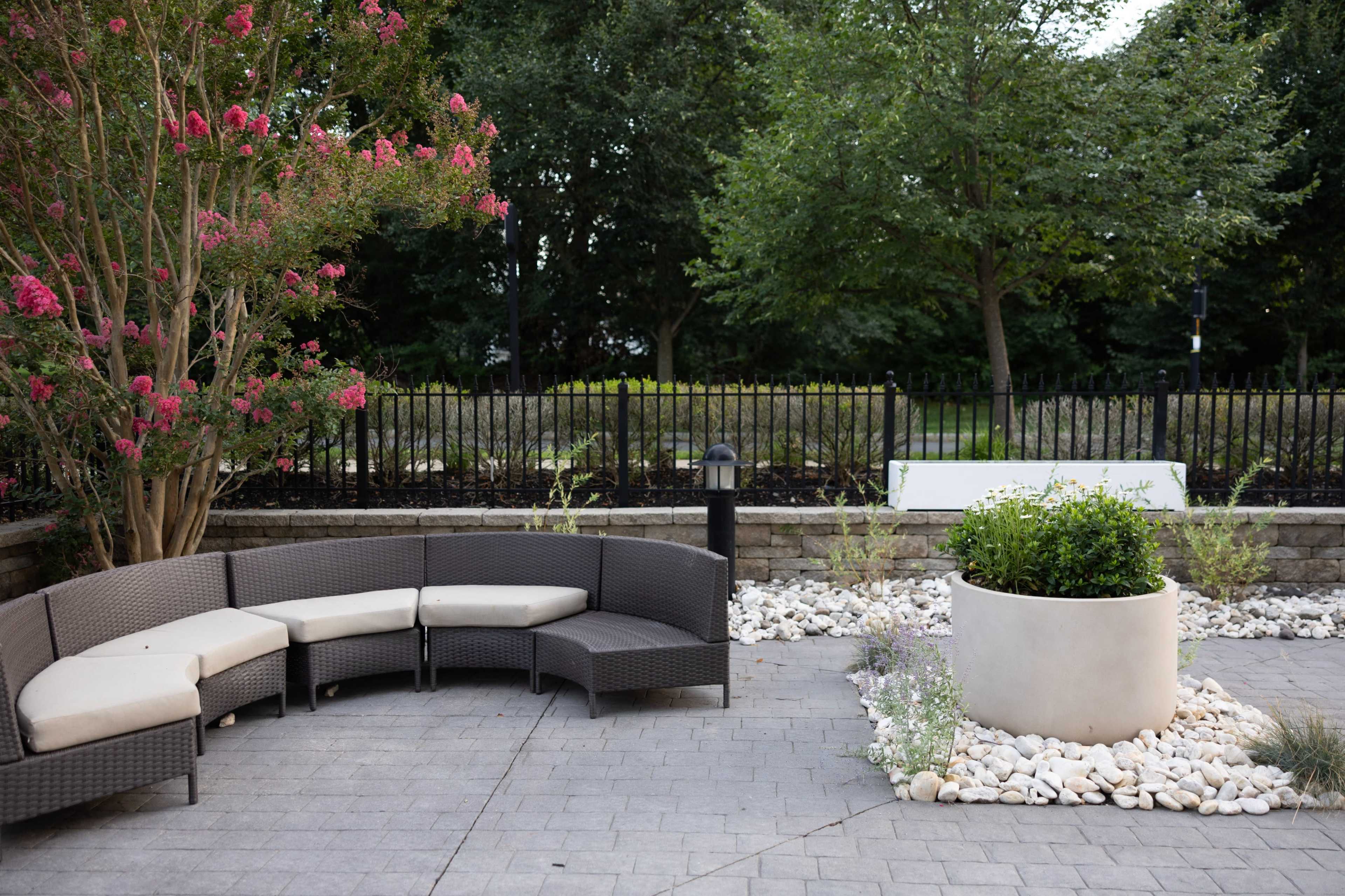 The image shows a patio area featuring a curved wicker seating arrangement, a large planter filled with plants, and landscaped stones, surrounded by trees and a black metal fence.