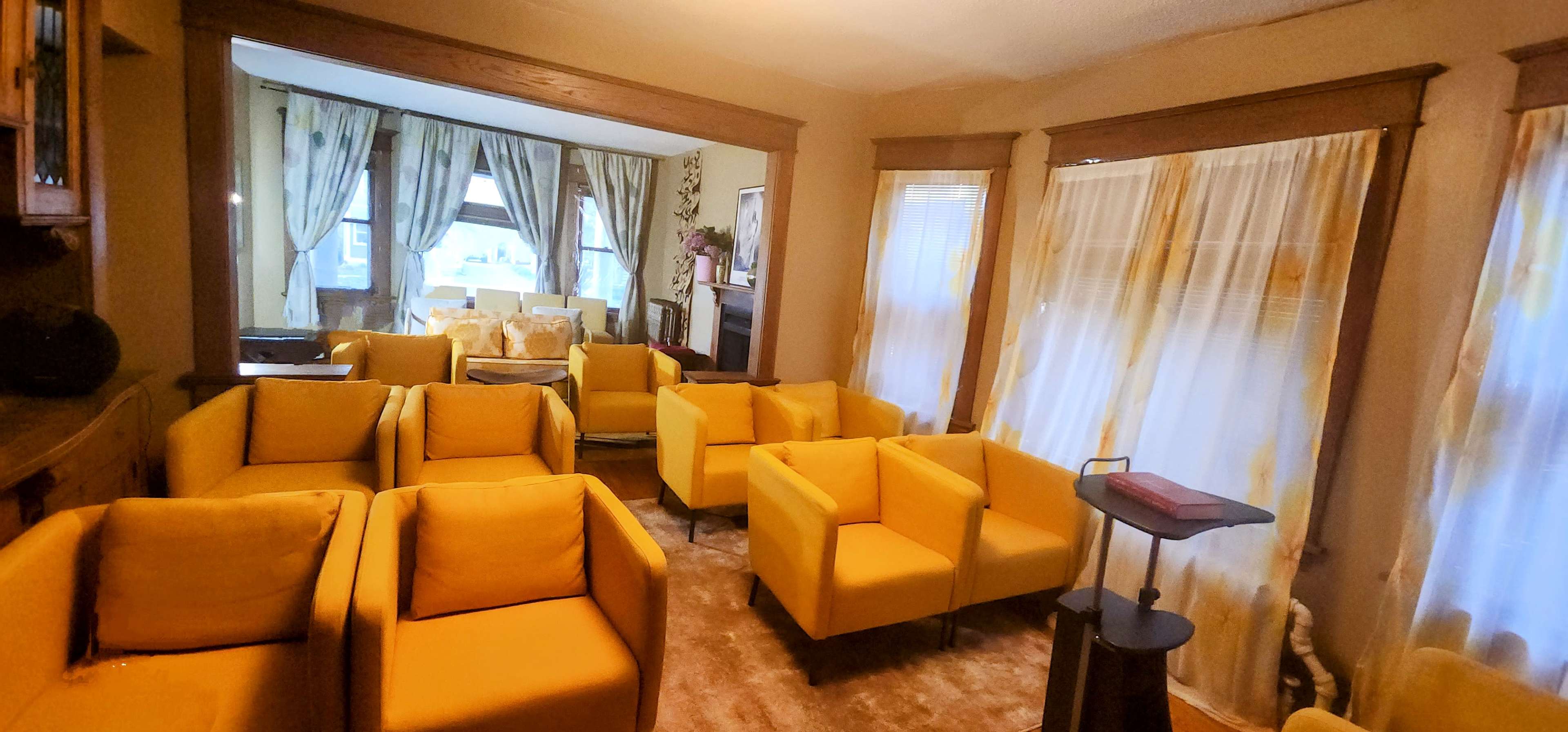 The image shows a living room furnished with several yellow armchairs arranged around a center table, with large windows and light curtains.