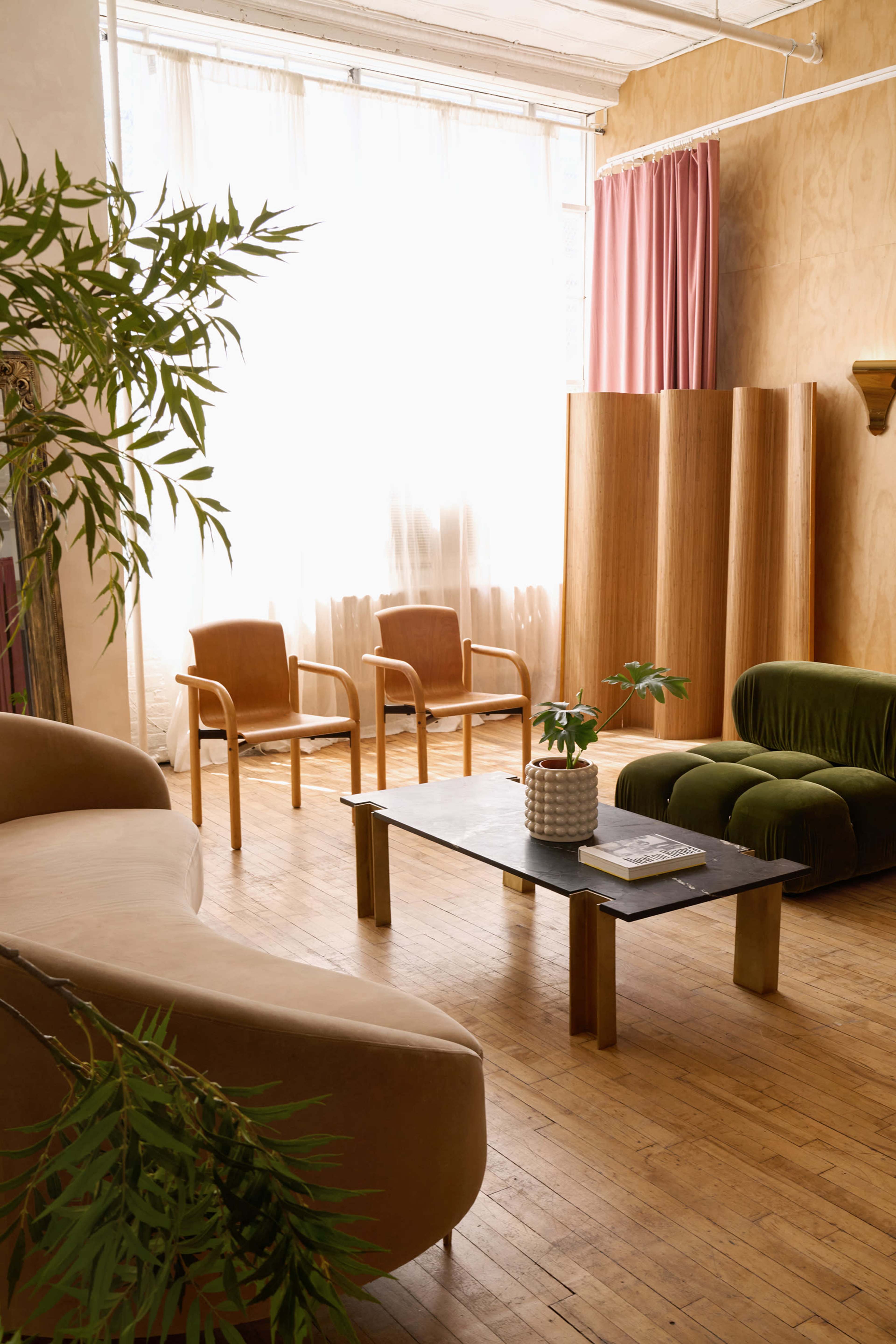 The image shows a stylish room with a mix of seating options, including a curved sofa, armchairs, and a coffee table, illuminated by natural light from a large window.