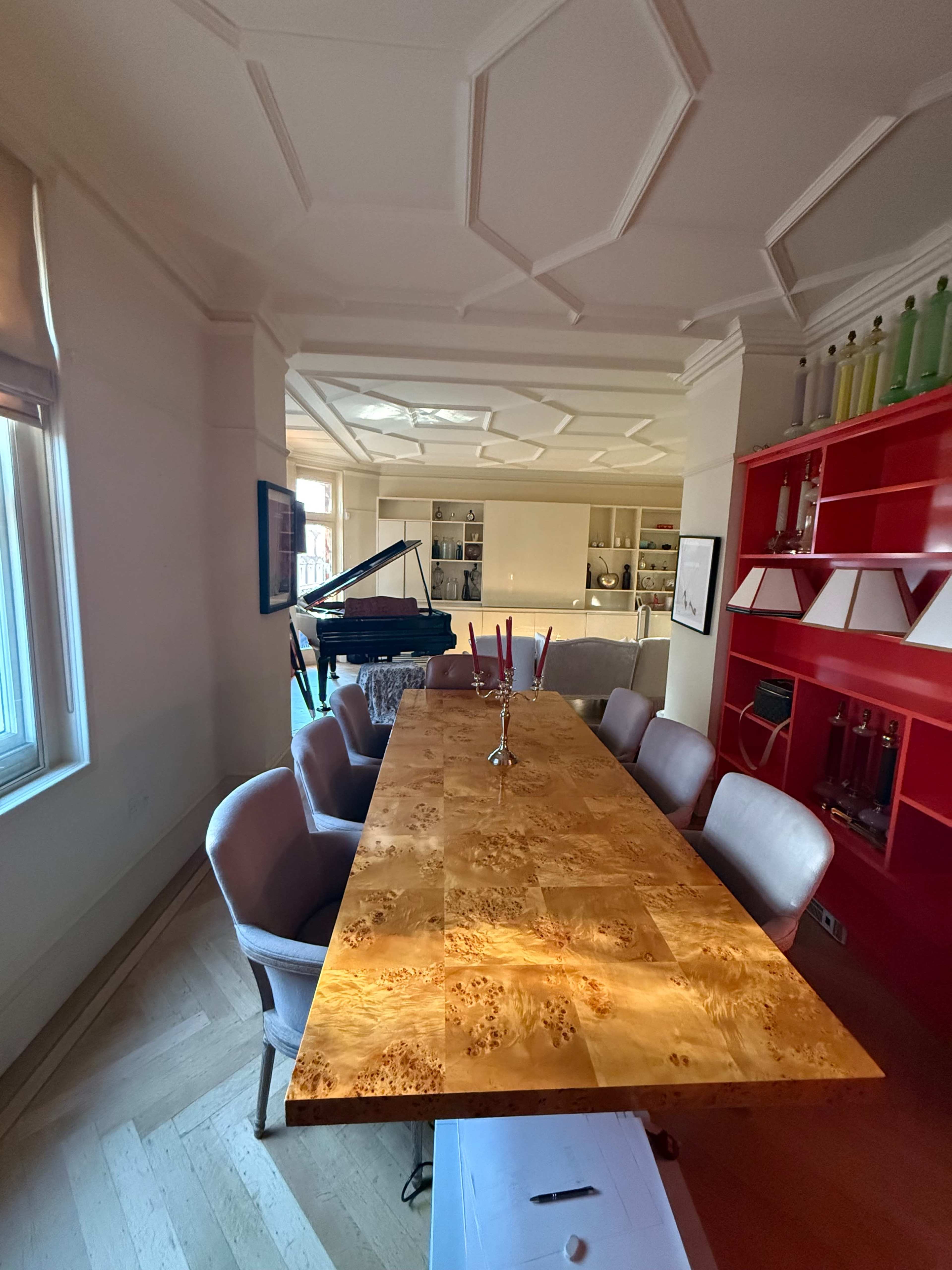 A spacious dining room features a long wooden table with a glossy finish surrounded by upholstered chairs, while a grand piano sits in the corner near shelves filled with glassware.