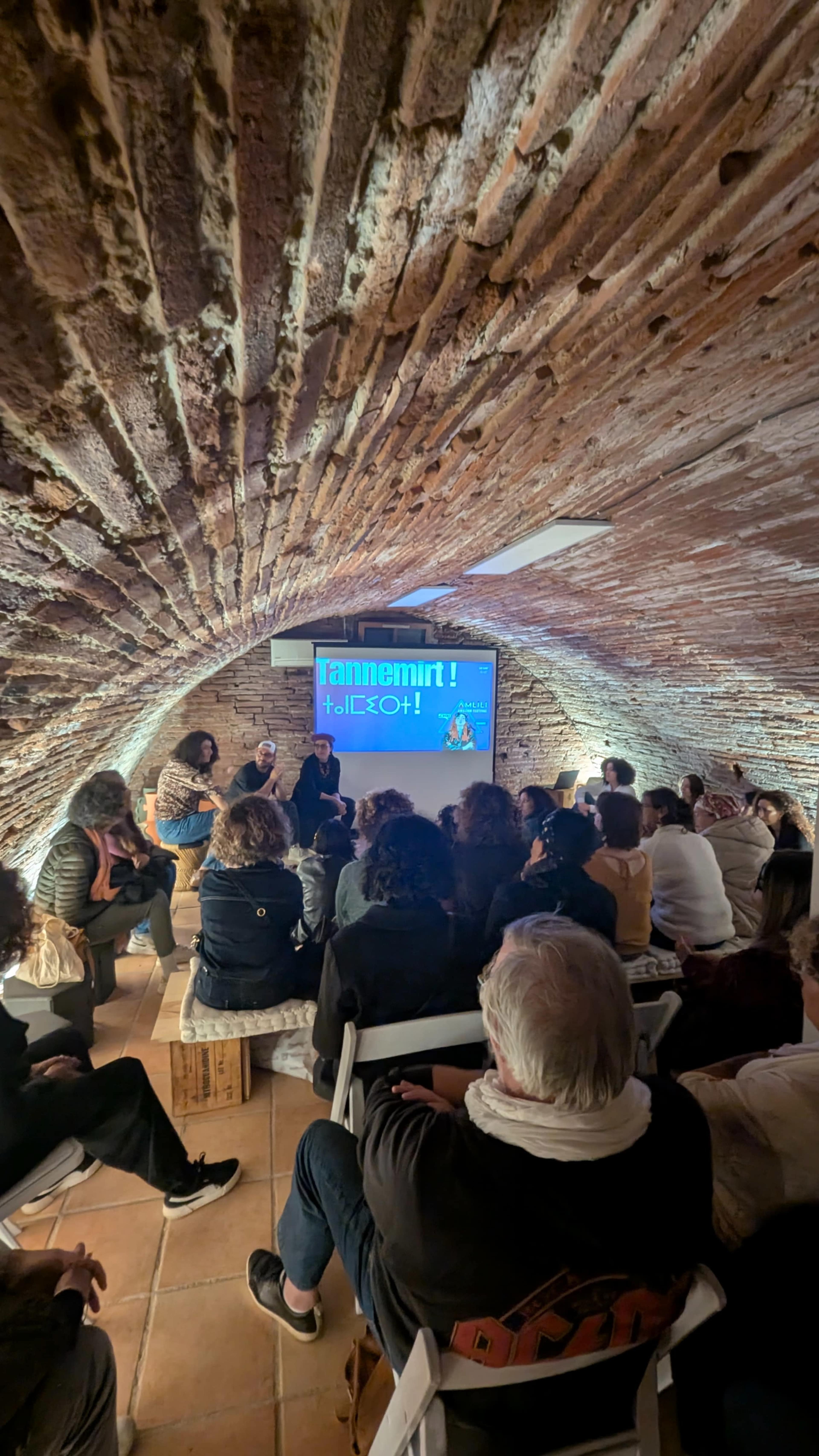 A group of people sits in a dimly lit, arched room while watching a presentation on a screen.