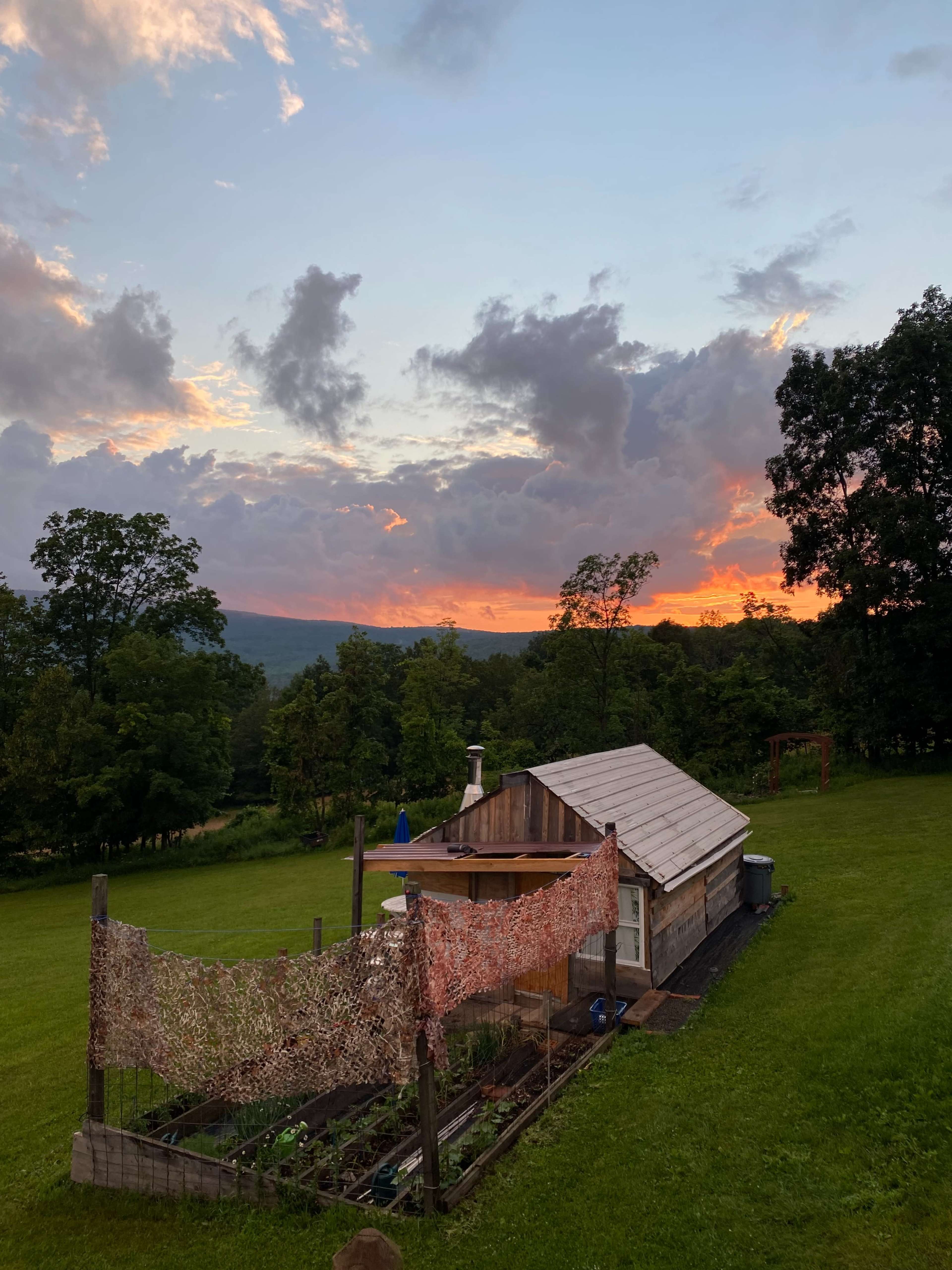 A small wooden cabin sits on green grass, surrounded by trees, with a colorful sunset and clouds overhead.