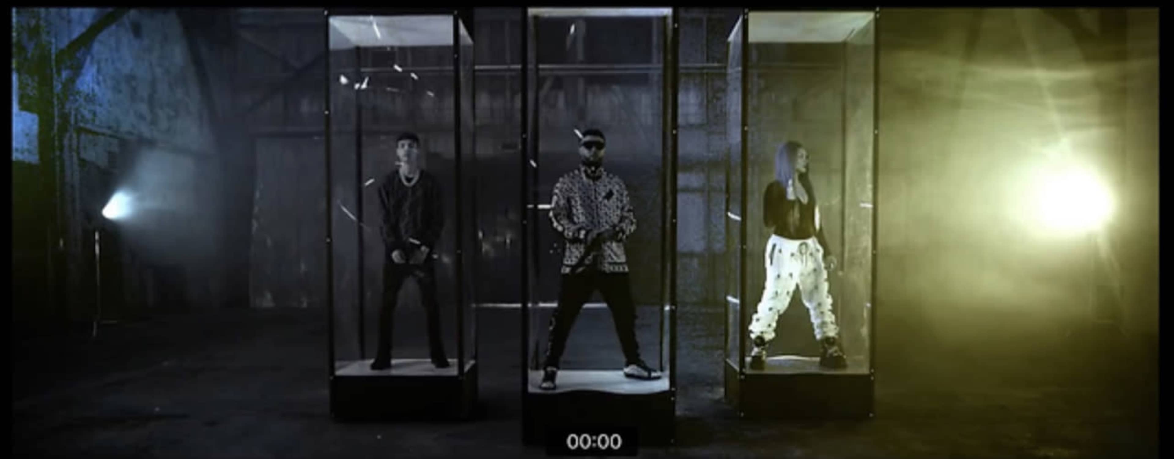 Three performers stand inside transparent glass boxes in a dimly lit industrial space.