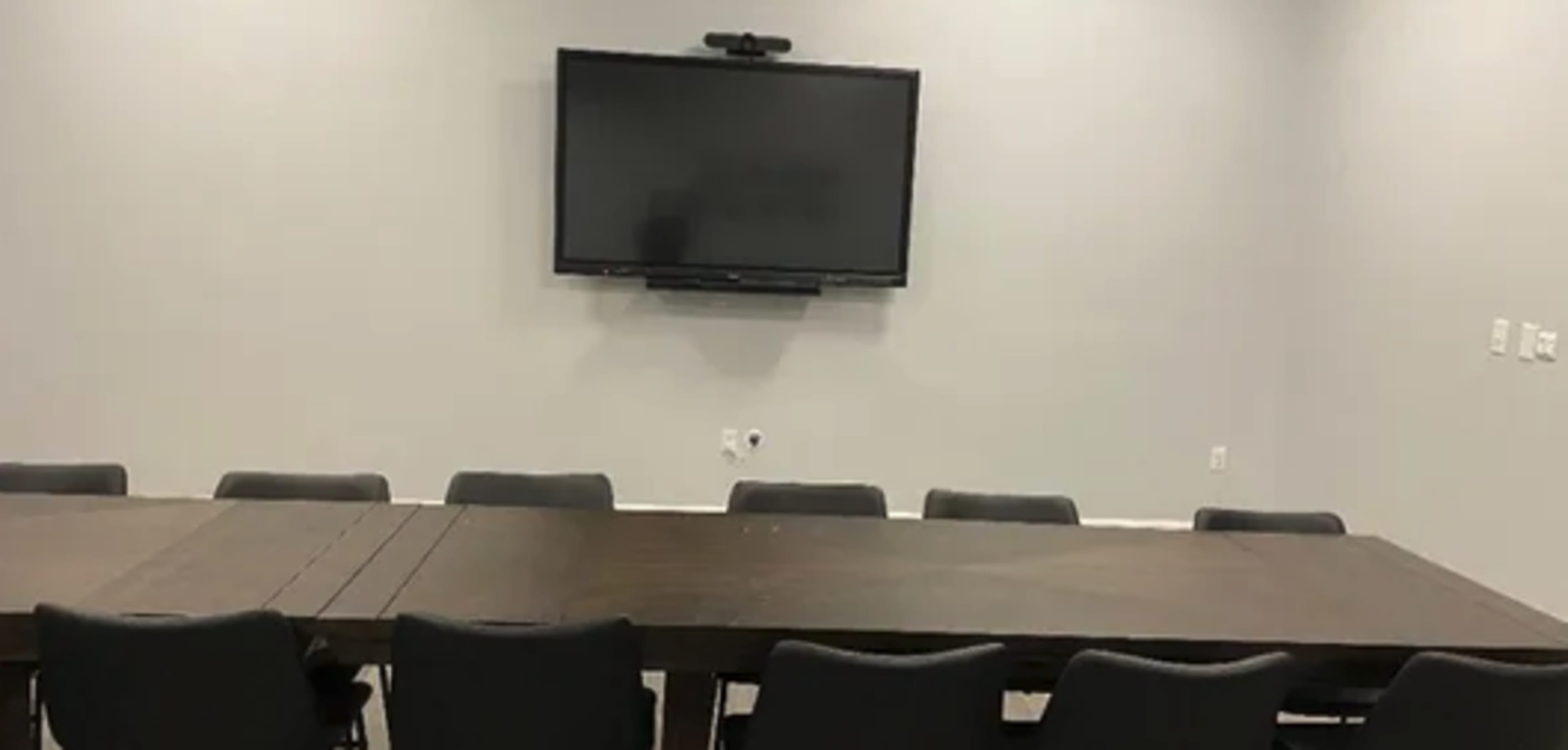 A conference room features a long wooden table surrounded by black chairs and a wall-mounted television.