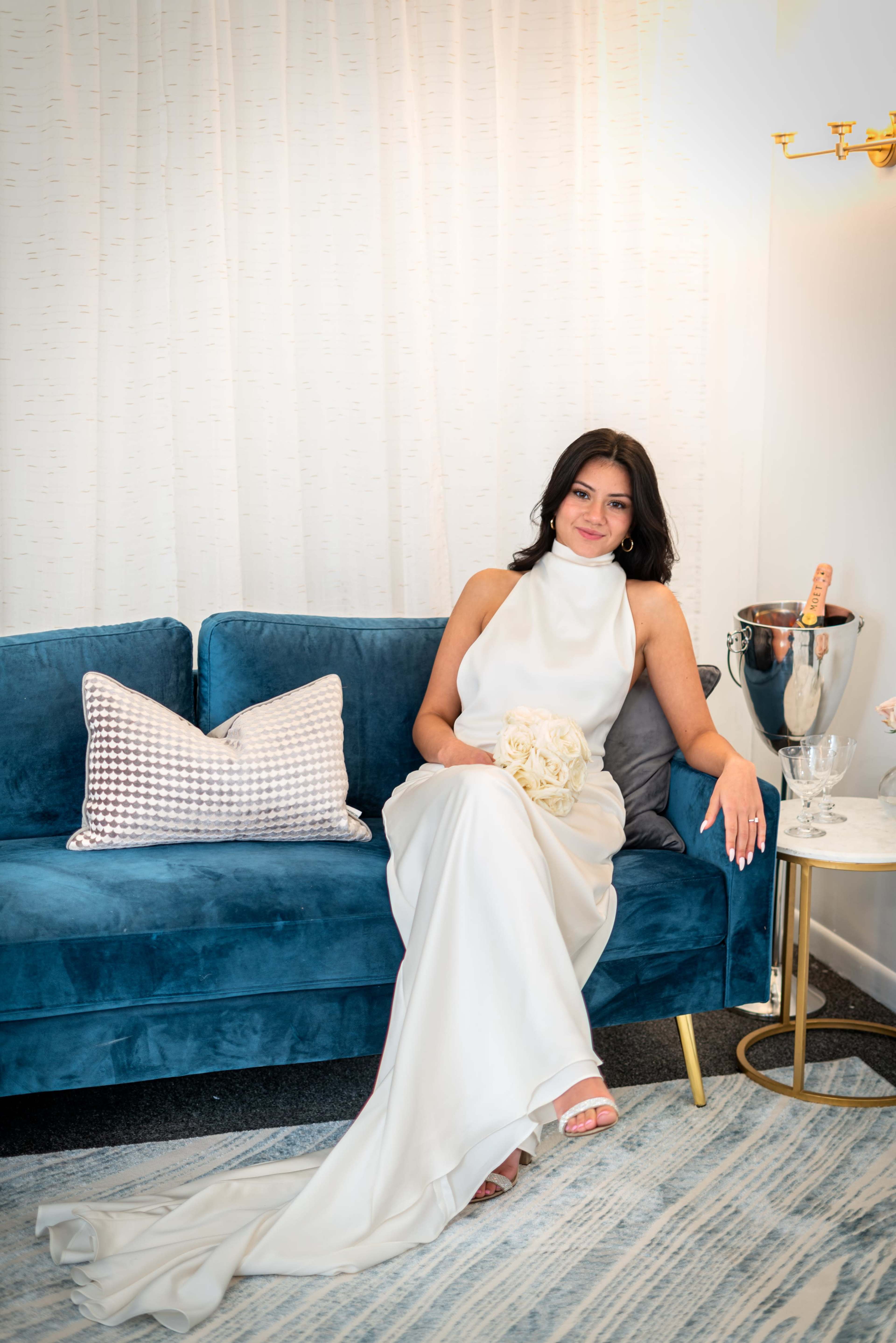 A woman in a white dress sits elegantly on a blue velvet sofa, holding a bouquet of flowers with a champagne bottle and glasses beside her.