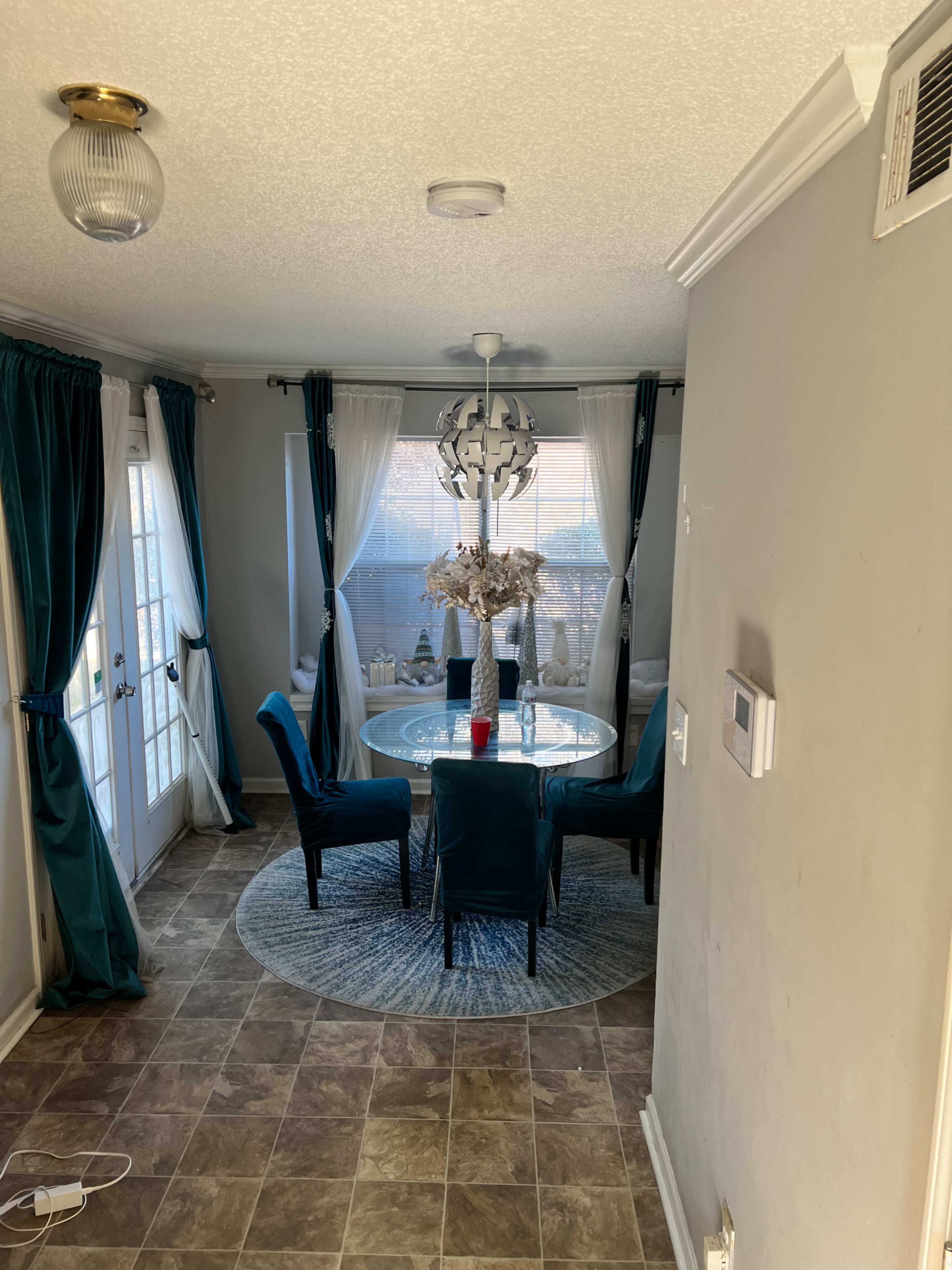 A dining area with a round glass table surrounded by four teal chairs, featuring a decorative light fixture and sheer curtains.