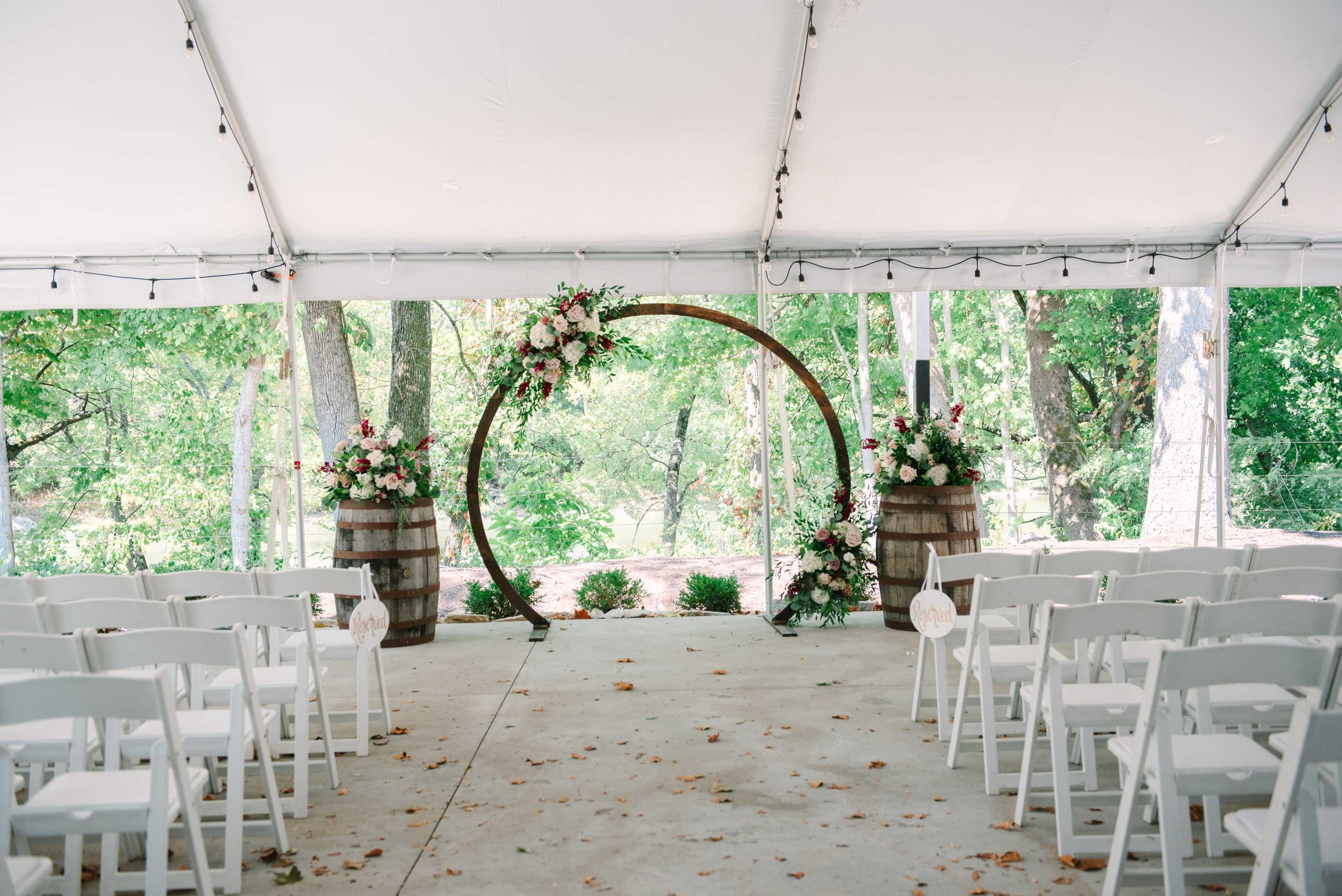 A wedding ceremony setup features a floral arch and wooden barrels surrounded by white chairs under a tent.
