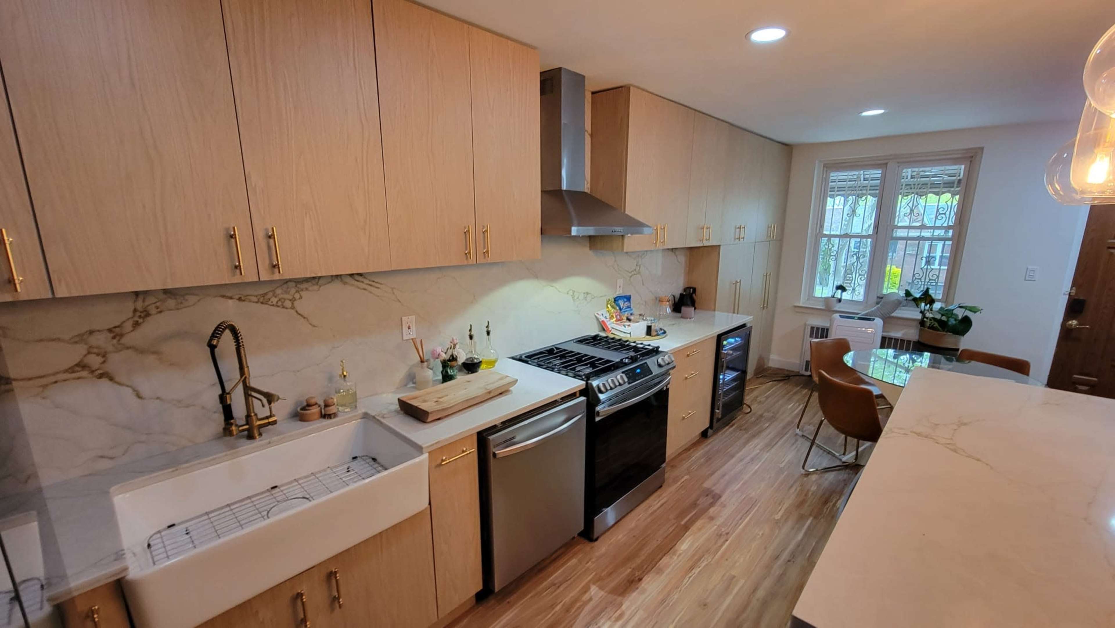 A modern kitchen featuring light wooden cabinets, a large farmhouse sink, stainless steel appliances, and marble countertops.