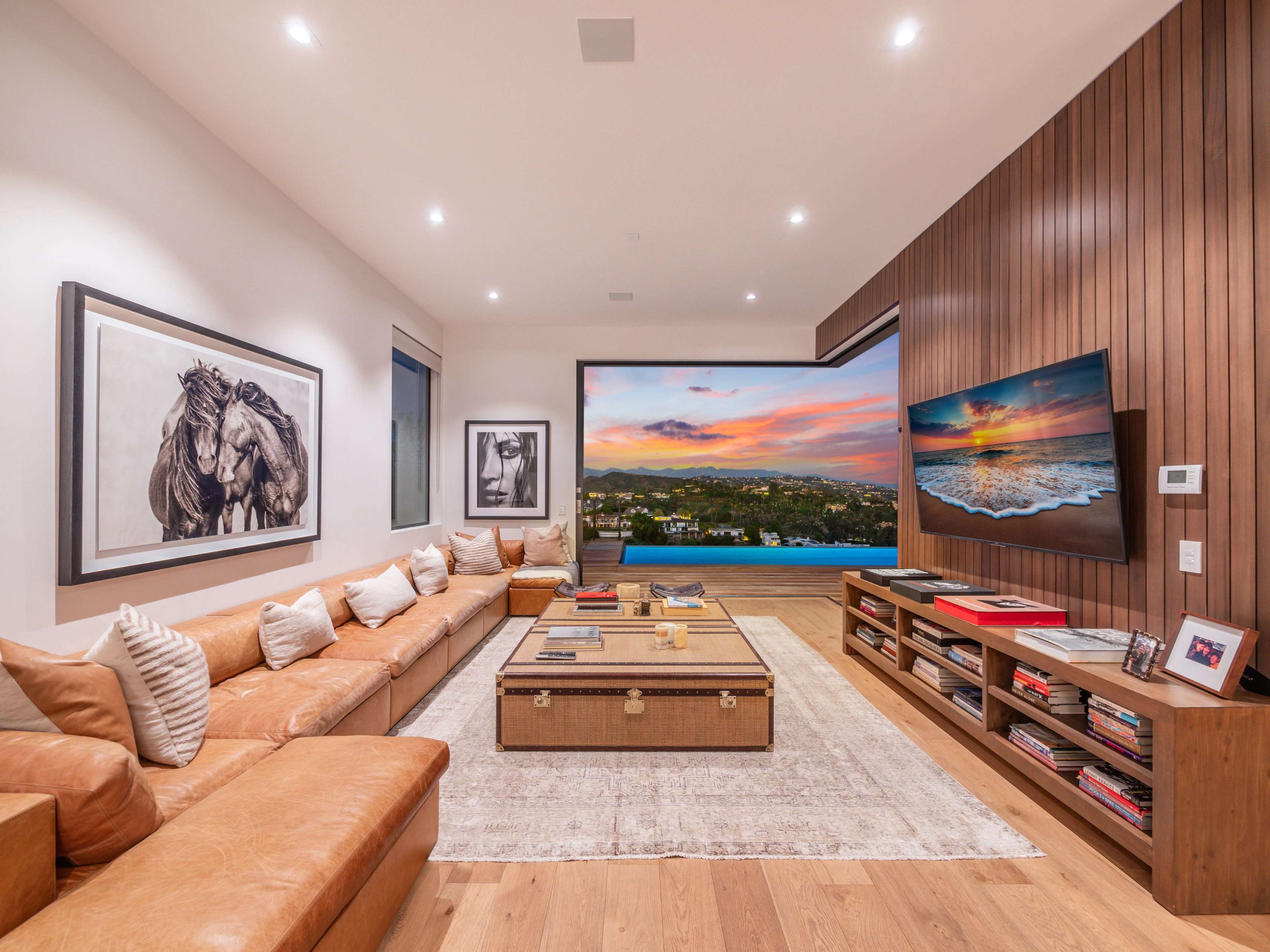 The living room features a large sectional sofa, a coffee table in the center, and a wall-mounted television displaying a sunset view.