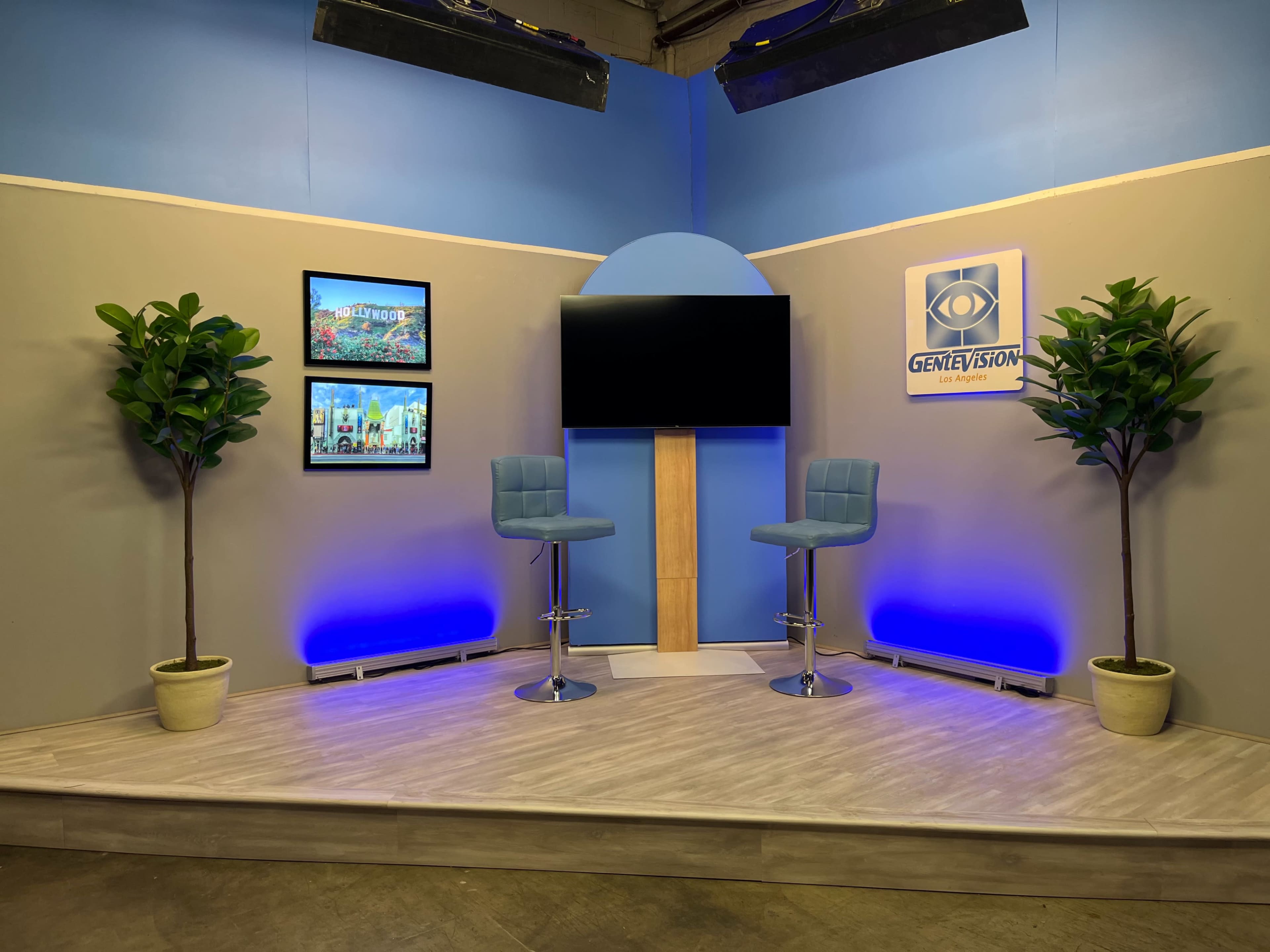 The image shows a television studio setup featuring two blue bar stools, a blank screen, and two decorative plants, with framed images mounted on the wall.