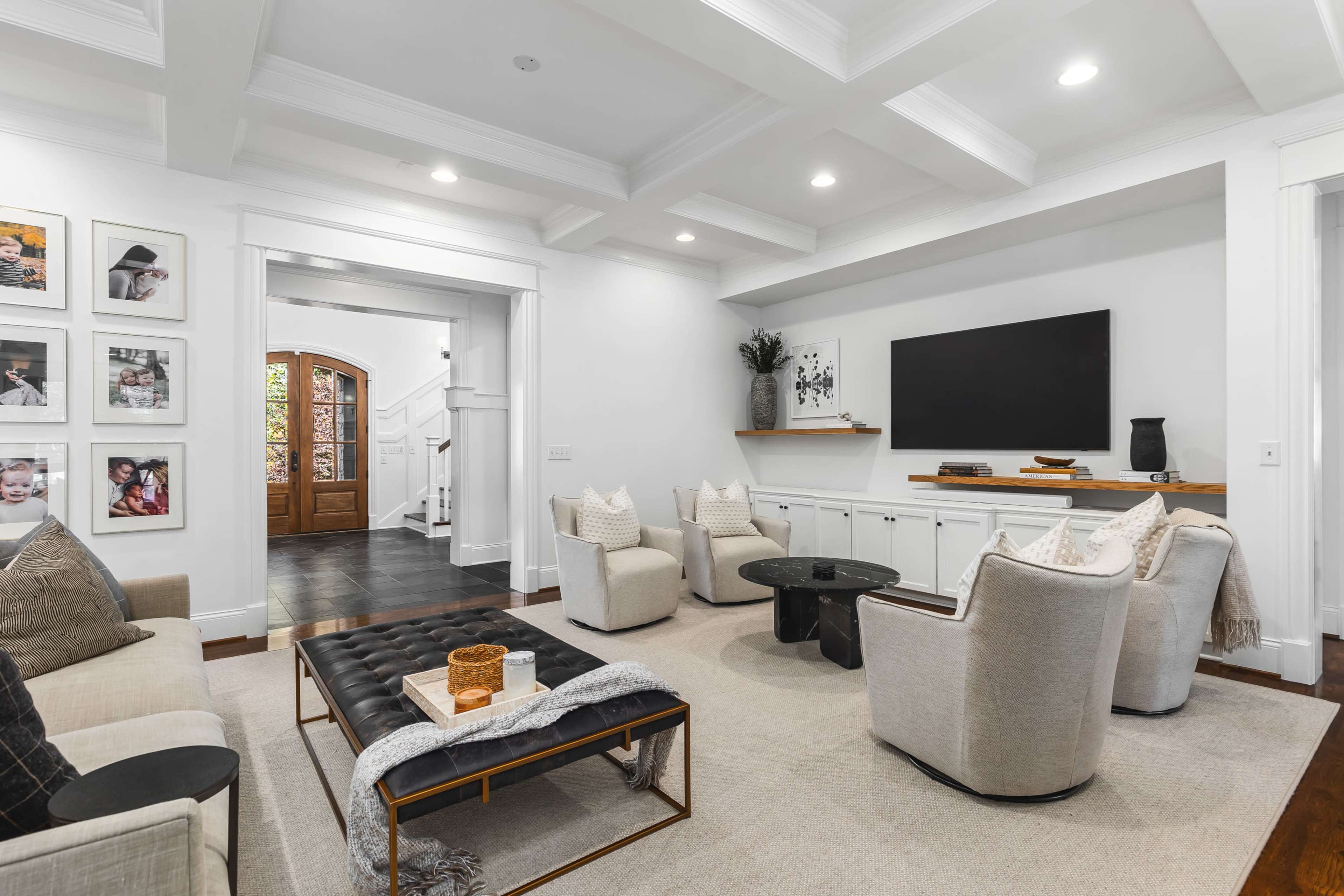 The image shows a modern living room with a light-colored furniture setup, a large television mounted on the wall, and decorative wall art framed in a gallery style.
