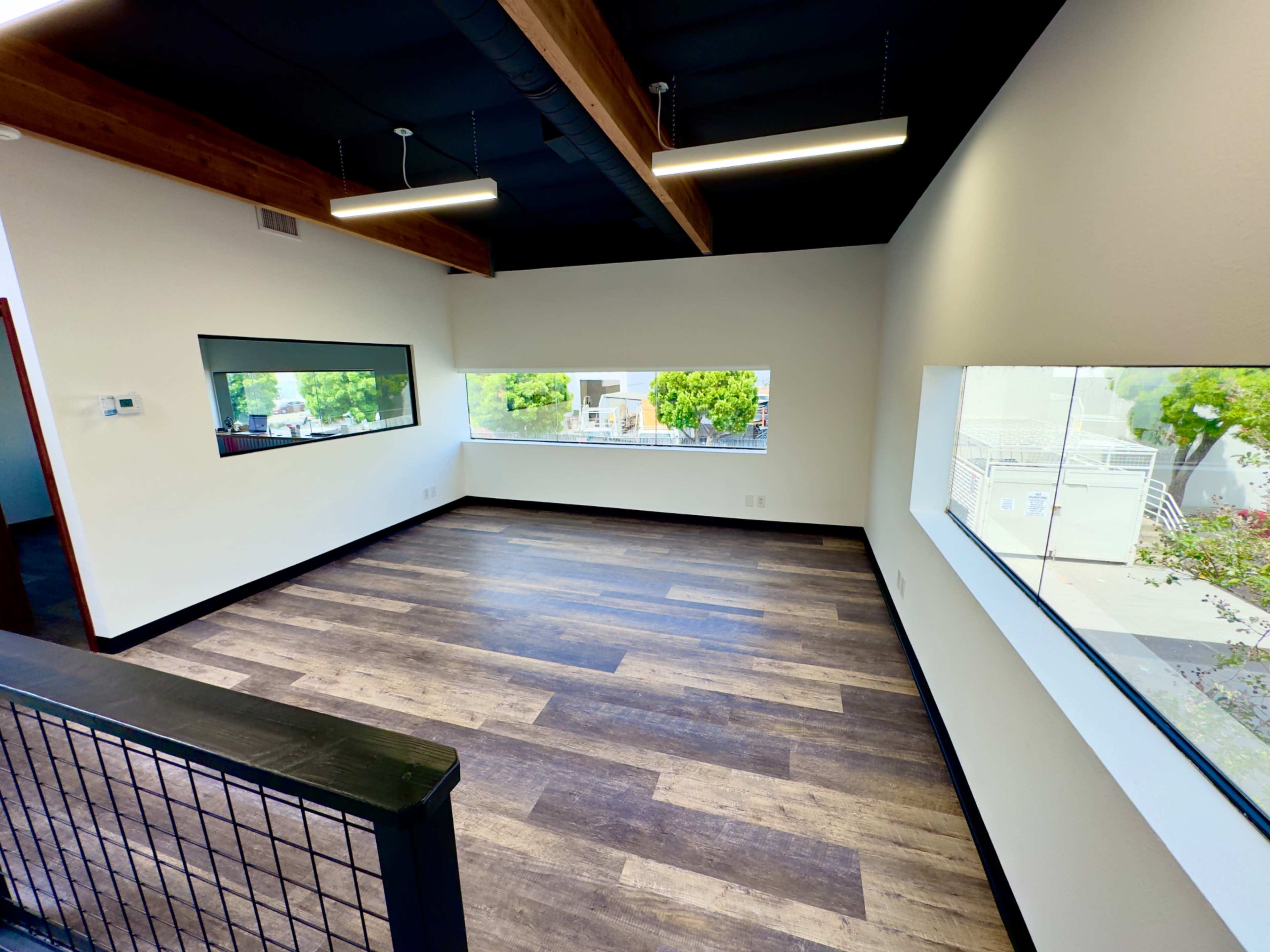 An empty room featuring large windows and wooden beams on the ceiling, with a wooden floor and white walls.