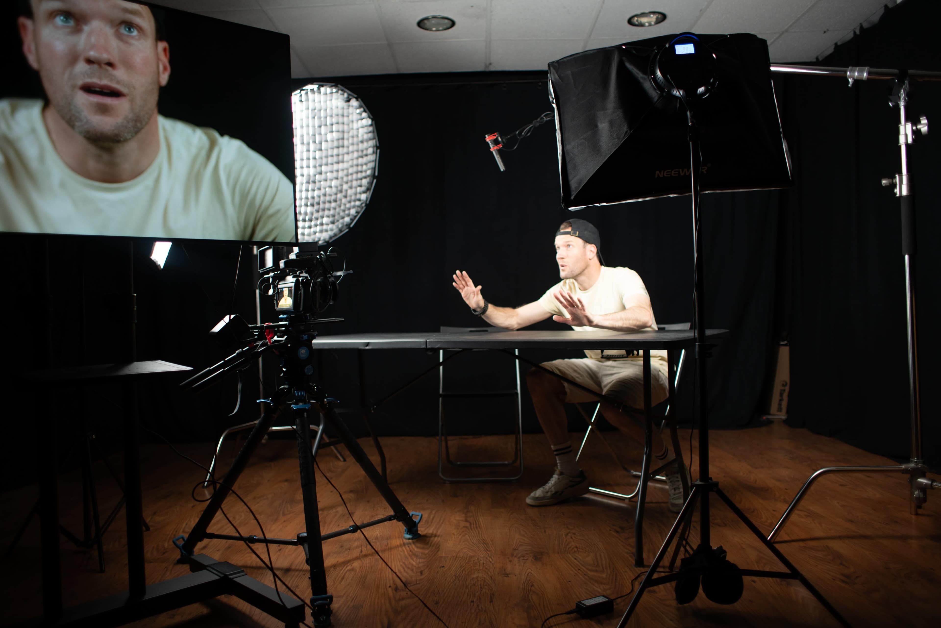 A person sits at a table in a dimly lit studio, gesturing while looking towards a camera and a large screen displaying their face.