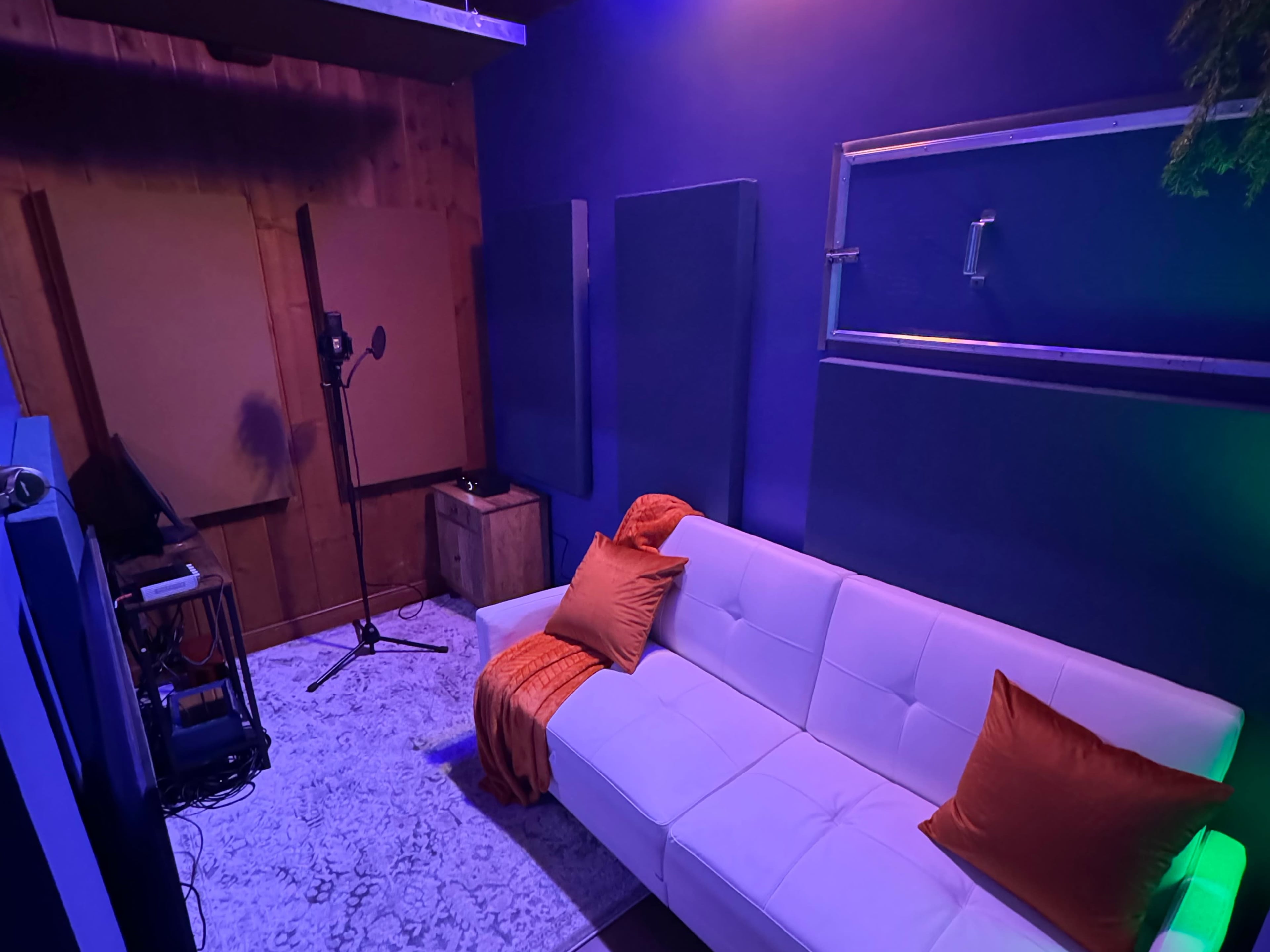 A cozy recording studio features a white sofa with orange pillows, acoustic panels on the walls, and a microphone stand positioned nearby.