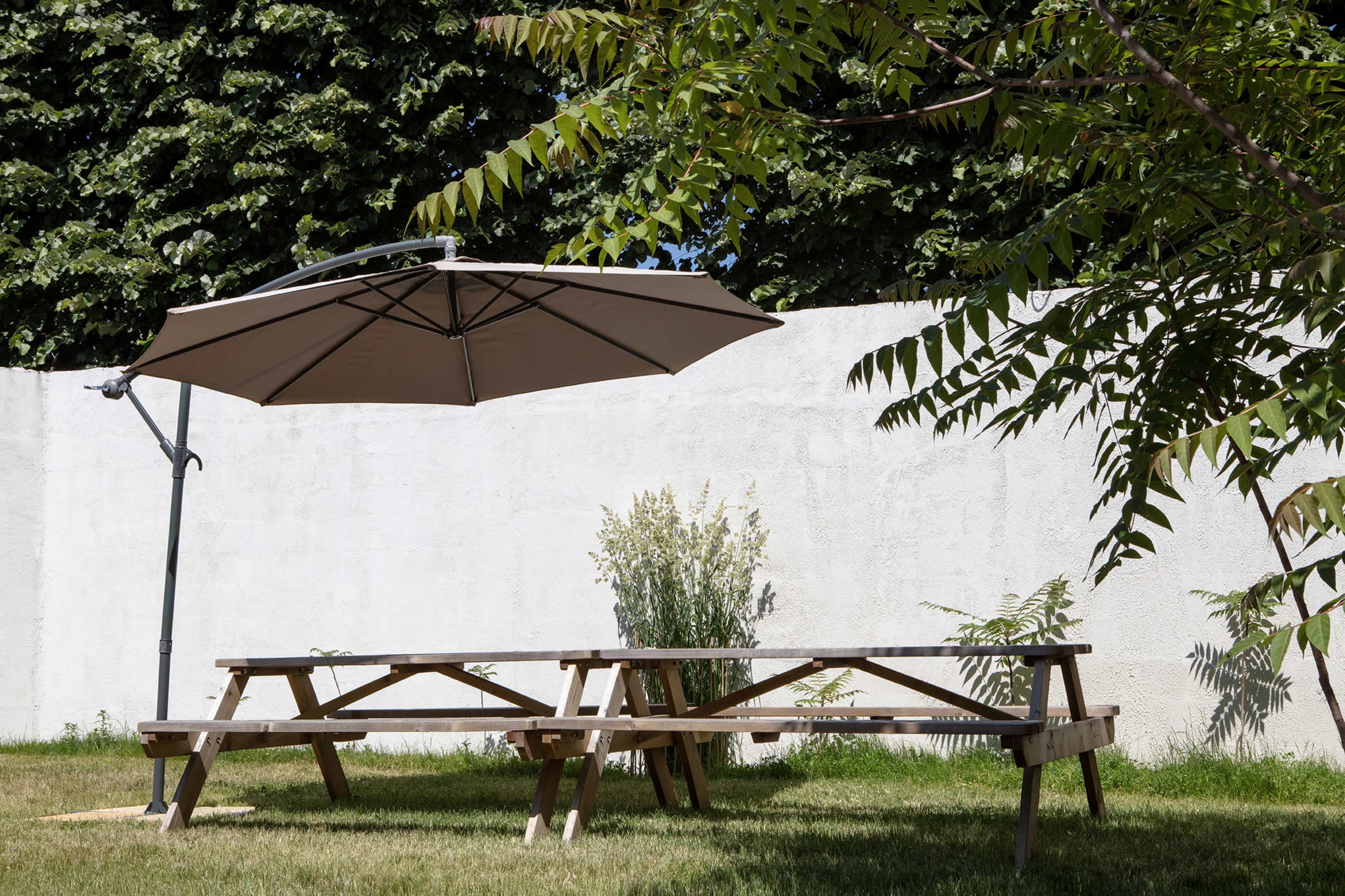 A wooden picnic table with a large umbrella is positioned on a grassy area next to a plain white wall and some greenery.