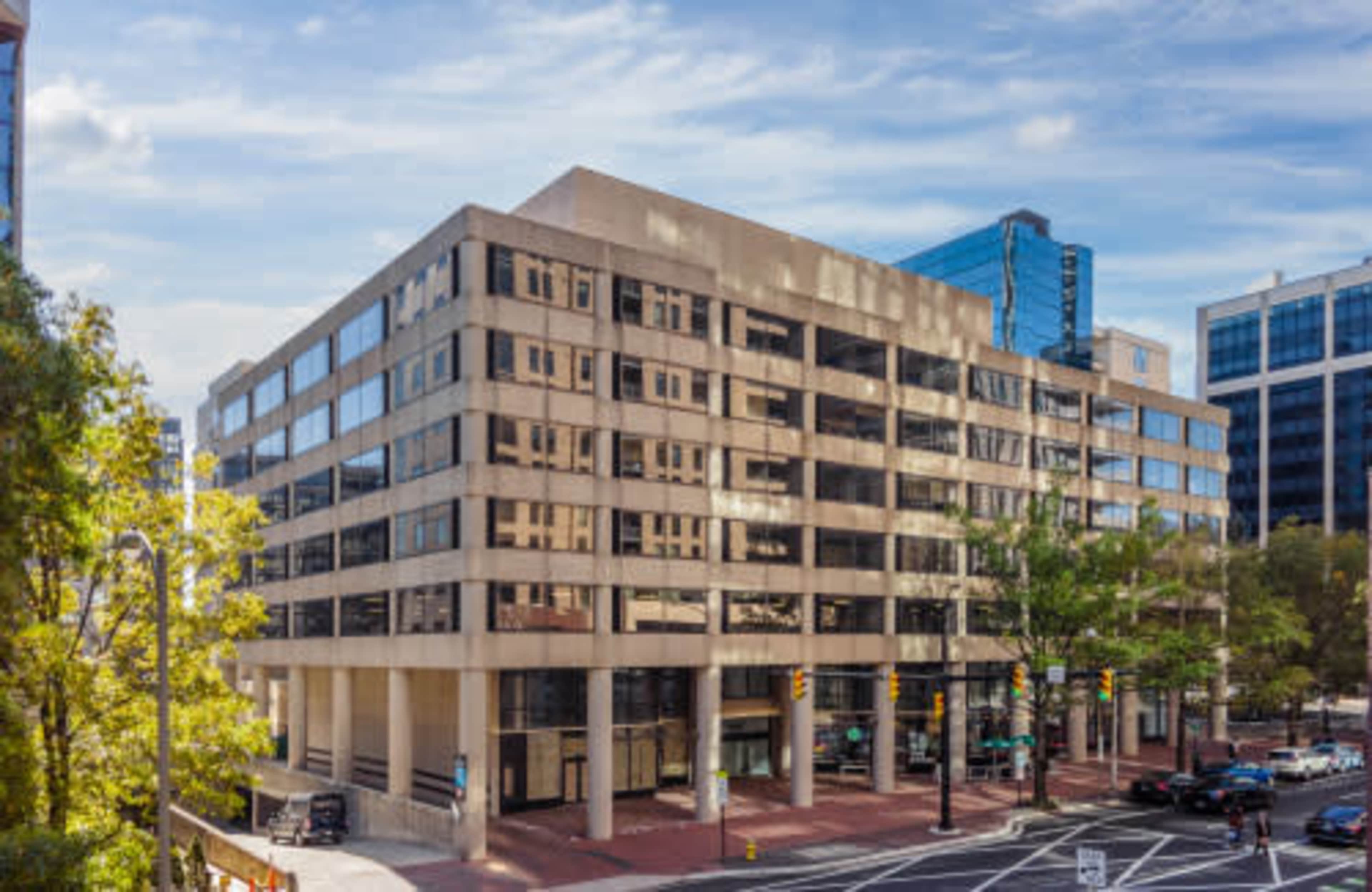 The image shows a modern, multi-story office building with large windows, located at an intersection surrounded by trees and other commercial structures.