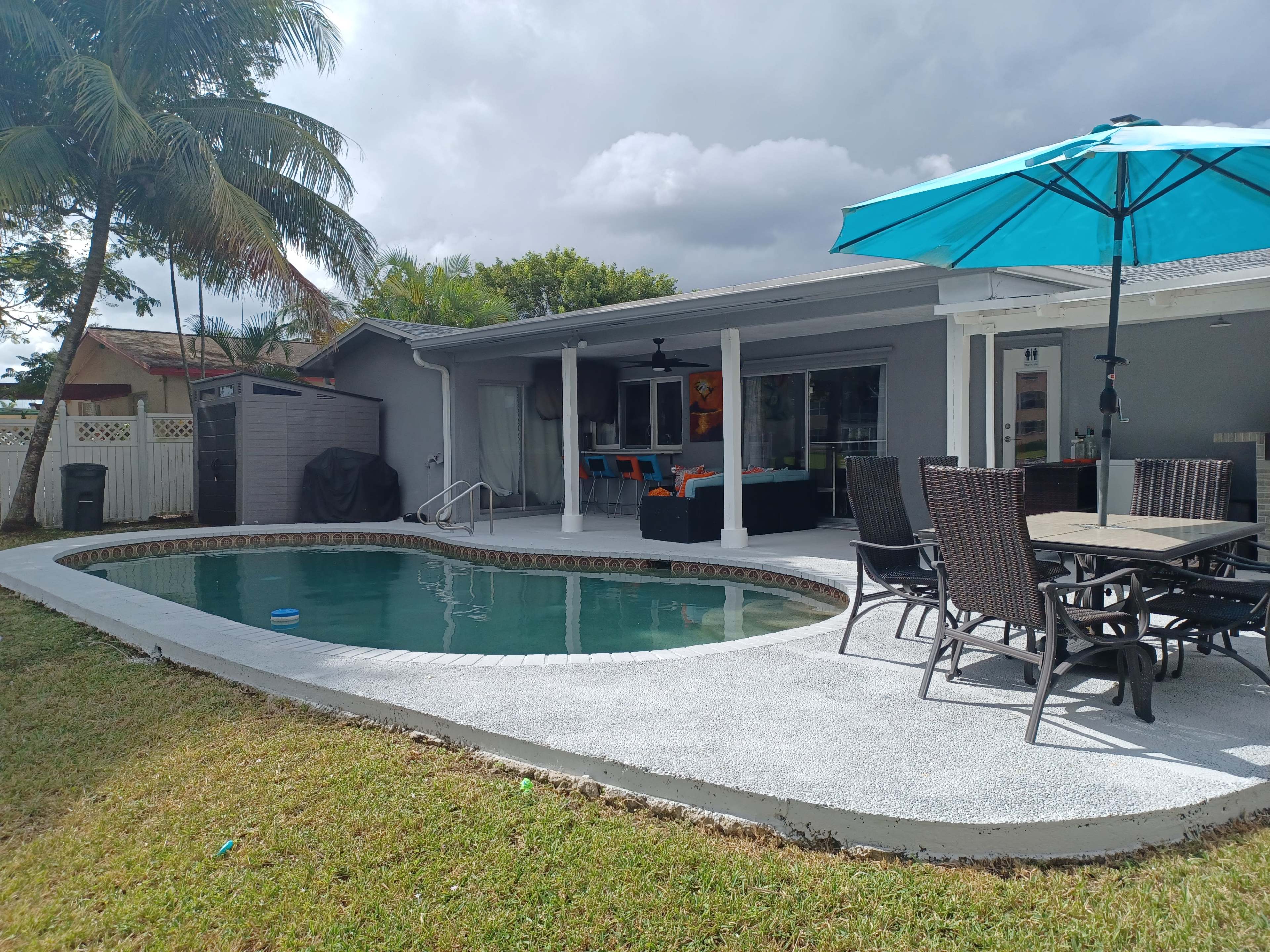 A backyard with a swimming pool, a dining table with chairs, and a large umbrella, surrounded by a landscaped area and a house in the background.