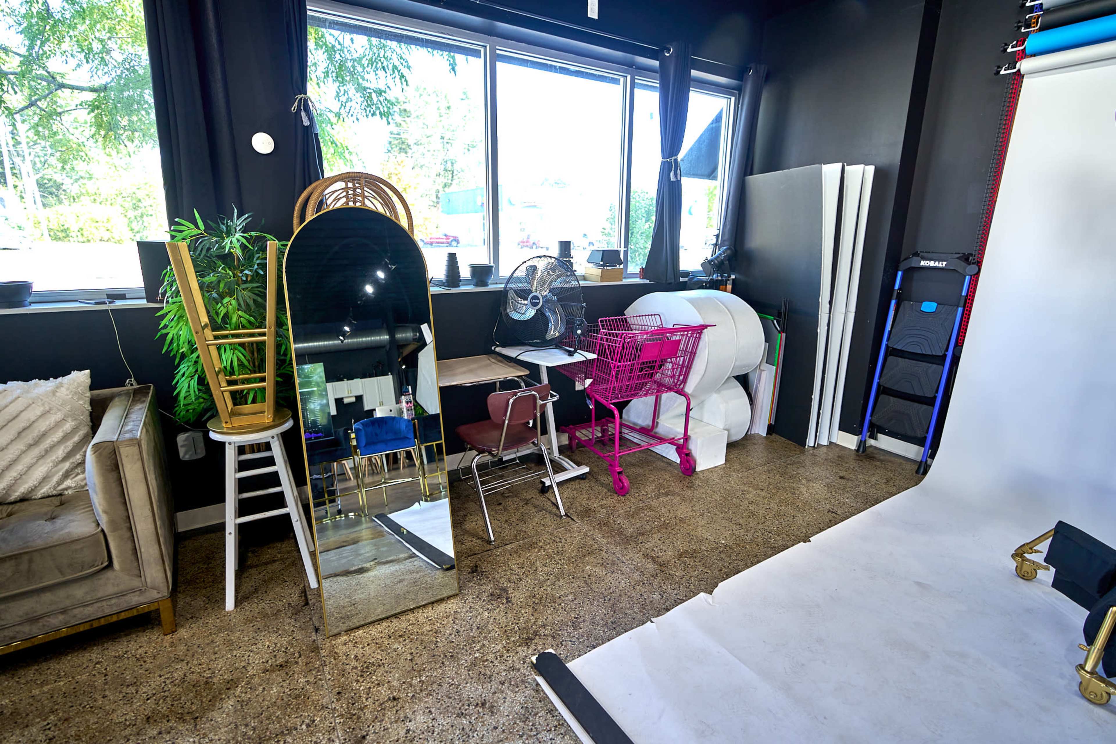 A photography studio features a mirror, a fan, chairs, a pink shopping cart, and various backgrounds beside a window.