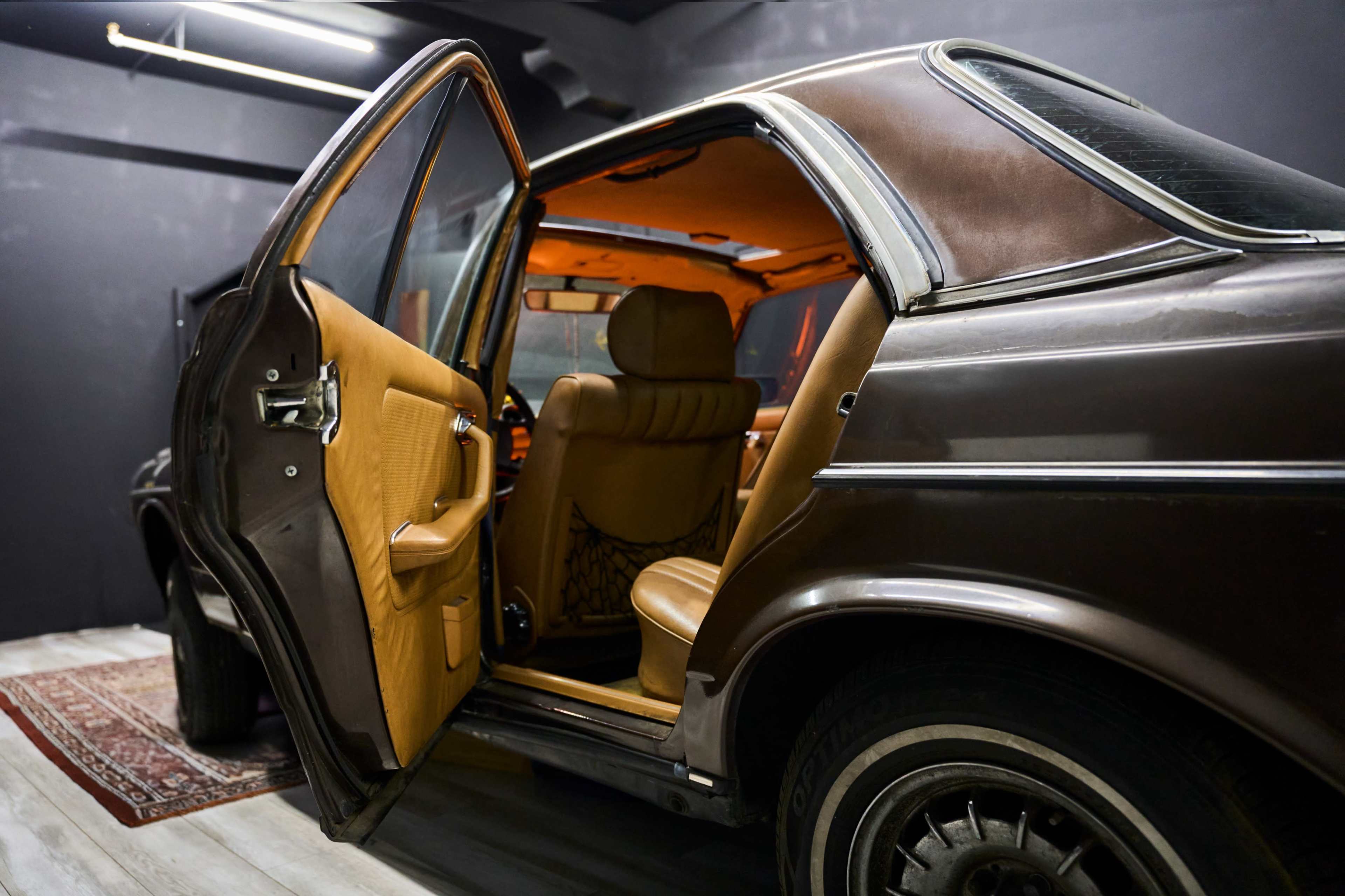 The image shows the interior of a vintage car with an open door, revealing tan leather seats and a patterned rug on the floor.