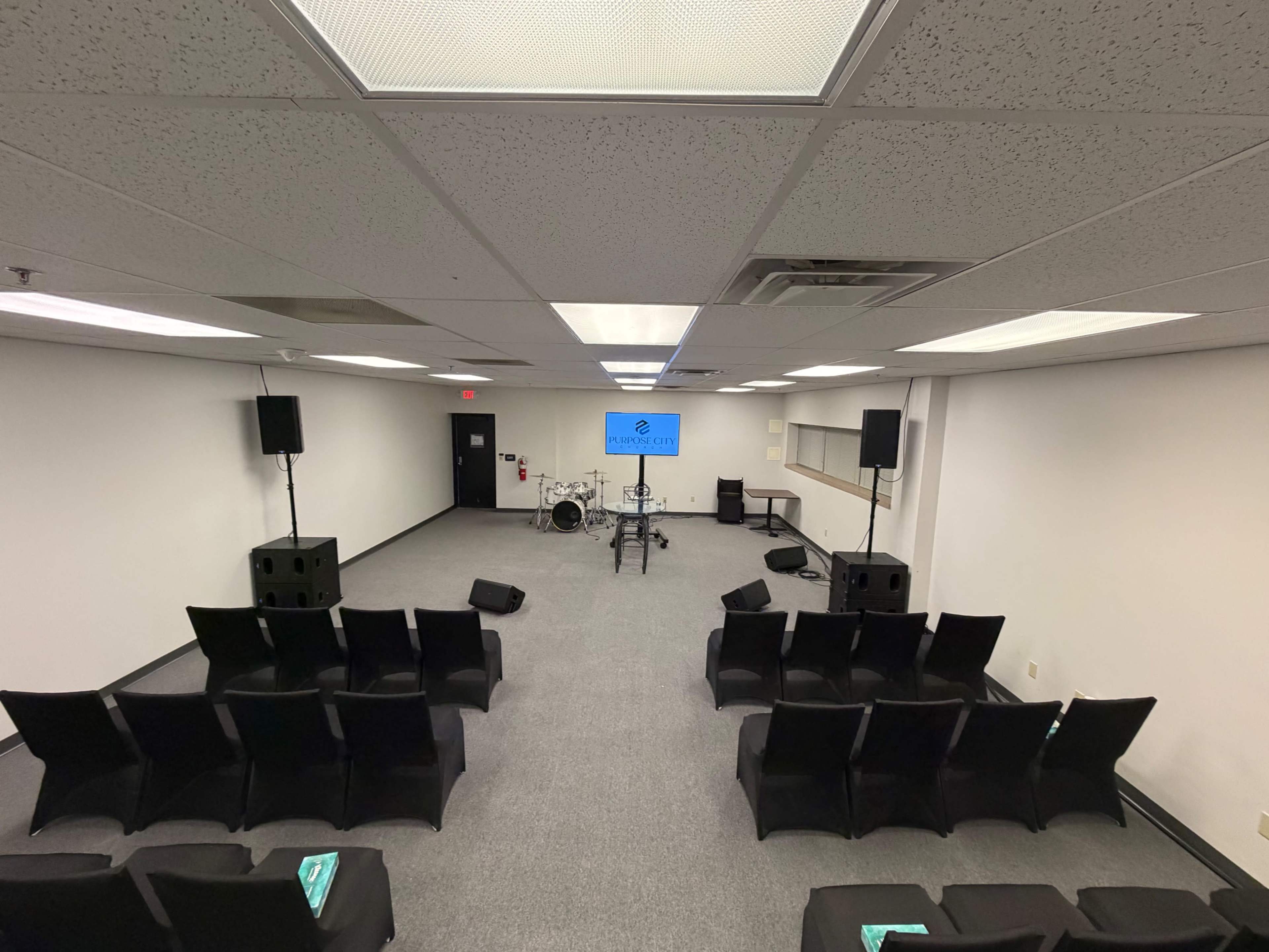 The image shows a large, empty room set up for an event, featuring rows of black chairs facing a stage with a drum set and a screen displaying a logo.