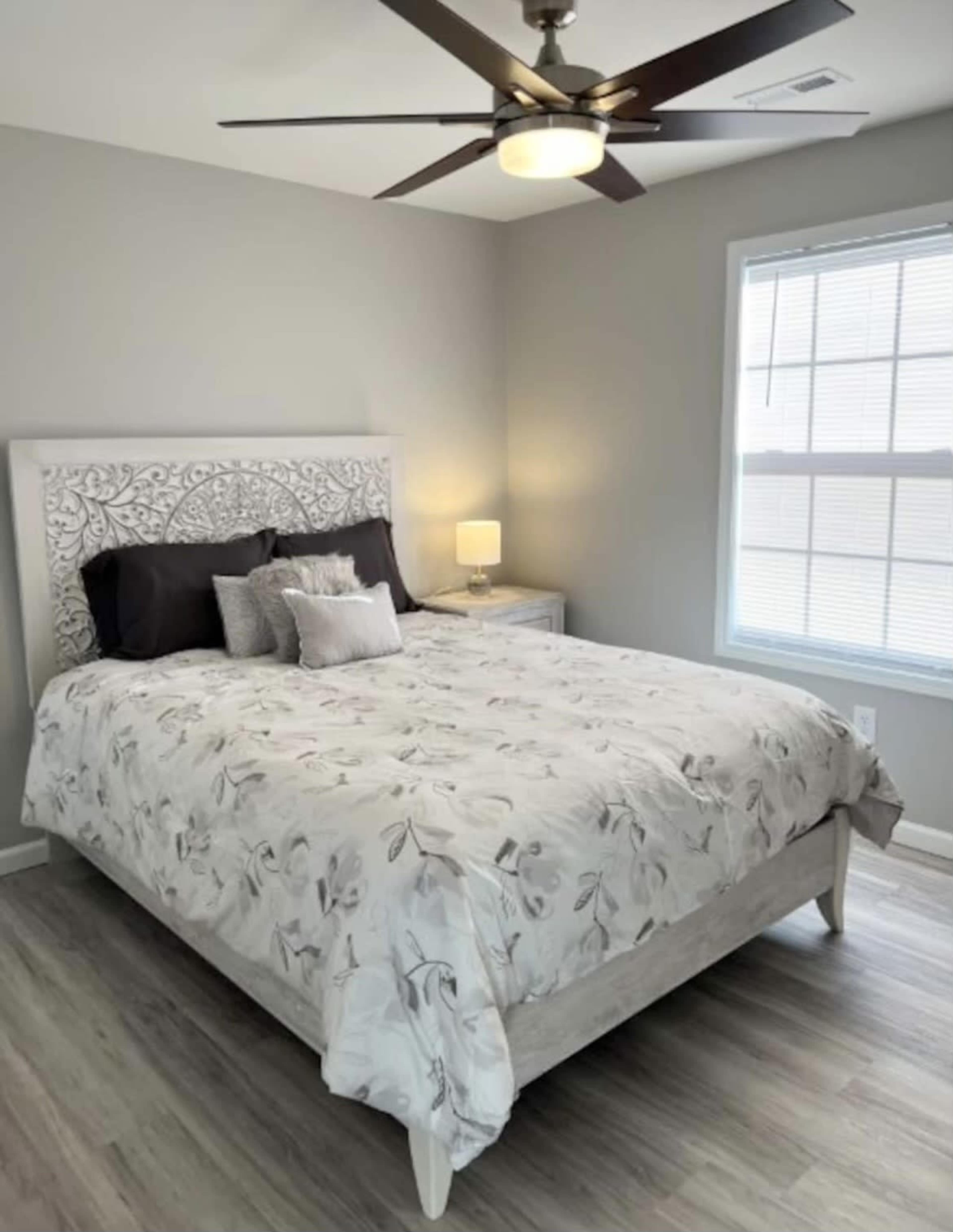 A modern bedroom with a queen-sized bed covered in a floral comforter, a nightstand with a lamp, and a window providing natural light.