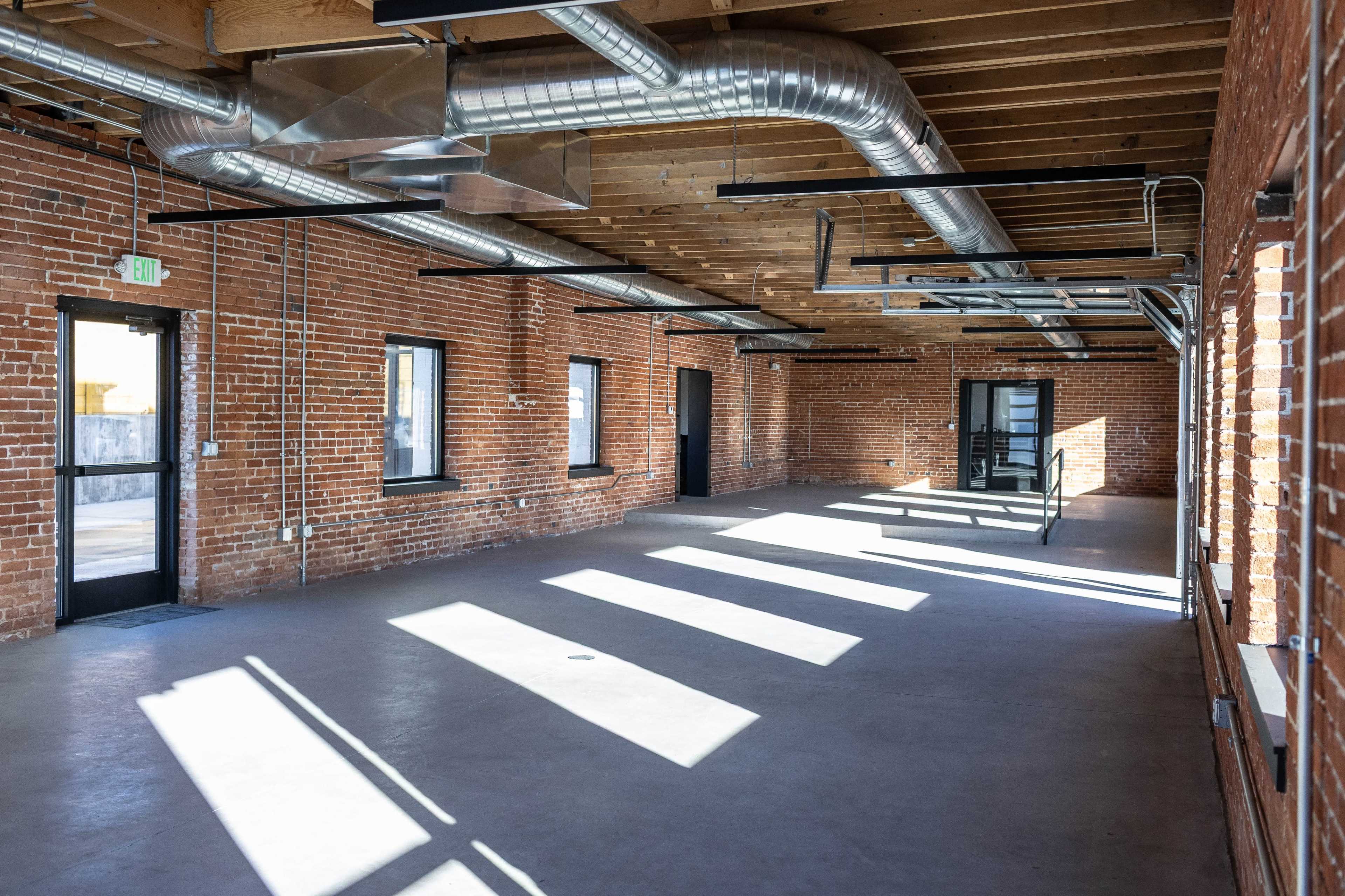 The image shows an empty industrial-style room with exposed brick walls, wooden beams, and large windows casting shadows on a concrete floor.