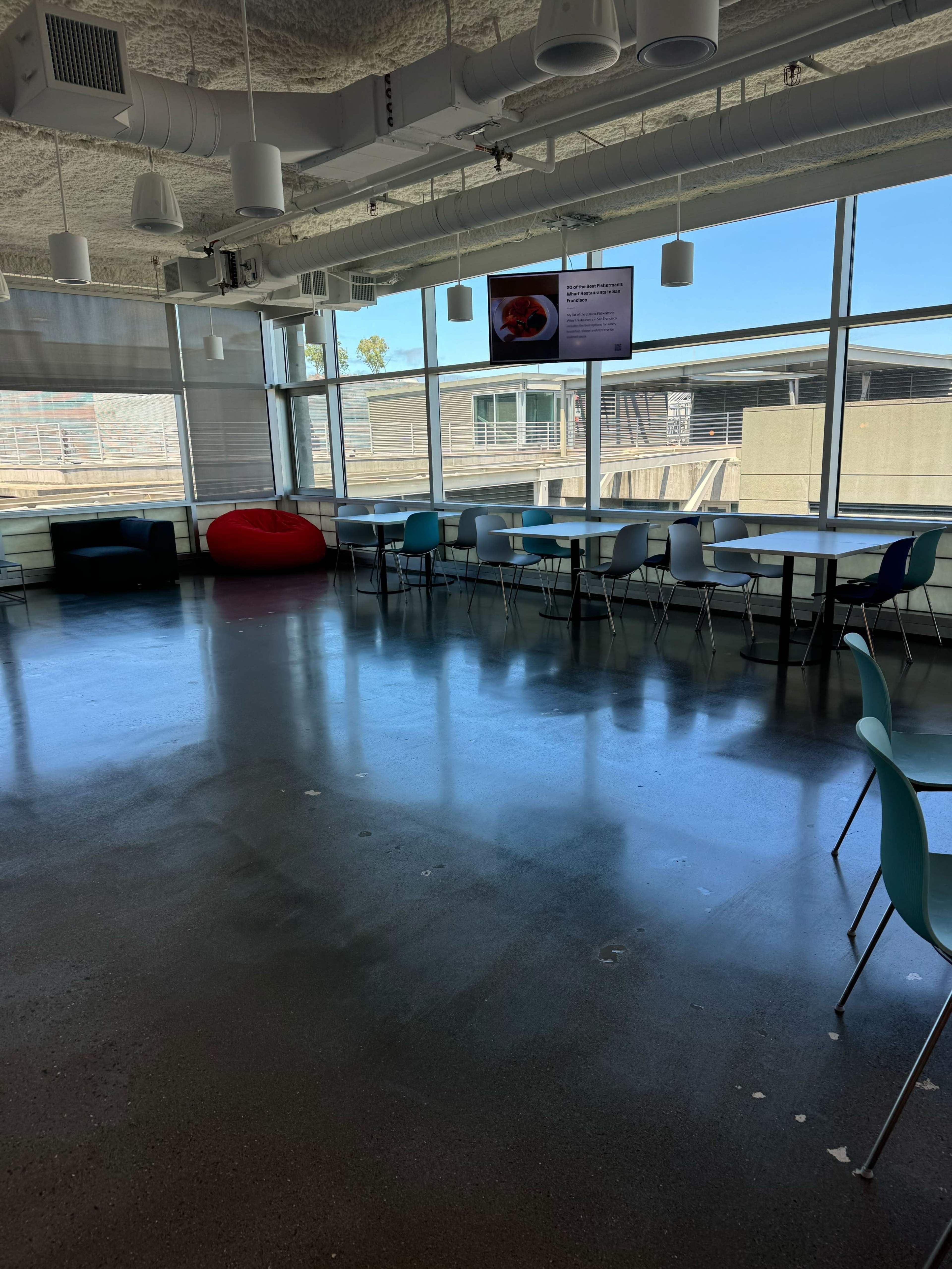 A modern, spacious break room with blue chairs, tables, and a television displaying information, all illuminated by natural light from large windows.